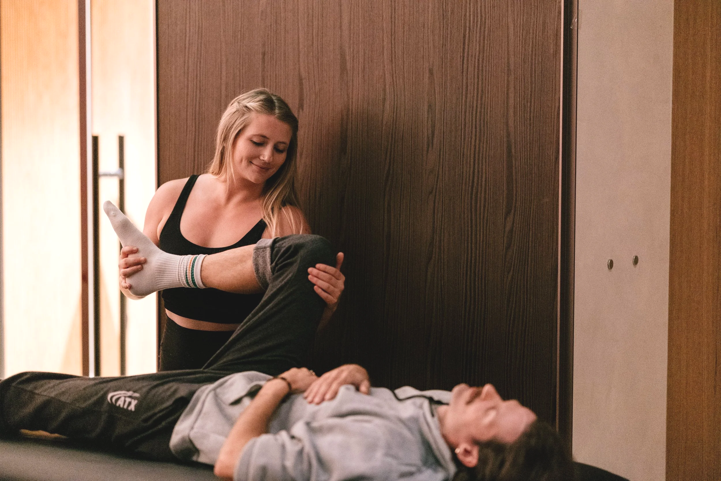 A woman appears to be helping a man who is lying on a bed, holding his knee, in a room with wooden paneling. She is doing stretch therapy and is assessing the internal rotation of his hip by moving his knee in and out.