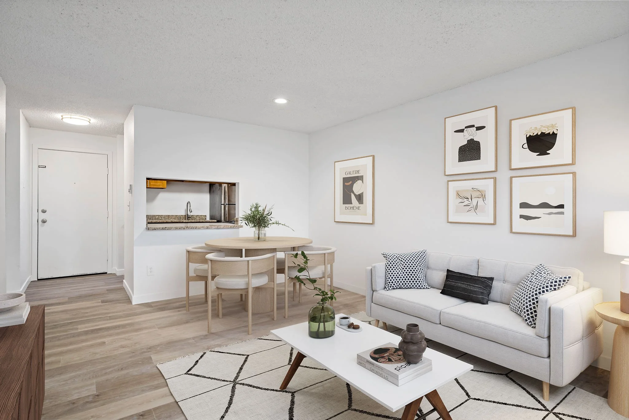 Living room with white walls, a white sofa with patterned pillows, a coffee table with books and vases, wall art, a small dining table with chairs, and a partial view of a kitchen with a small window opening.