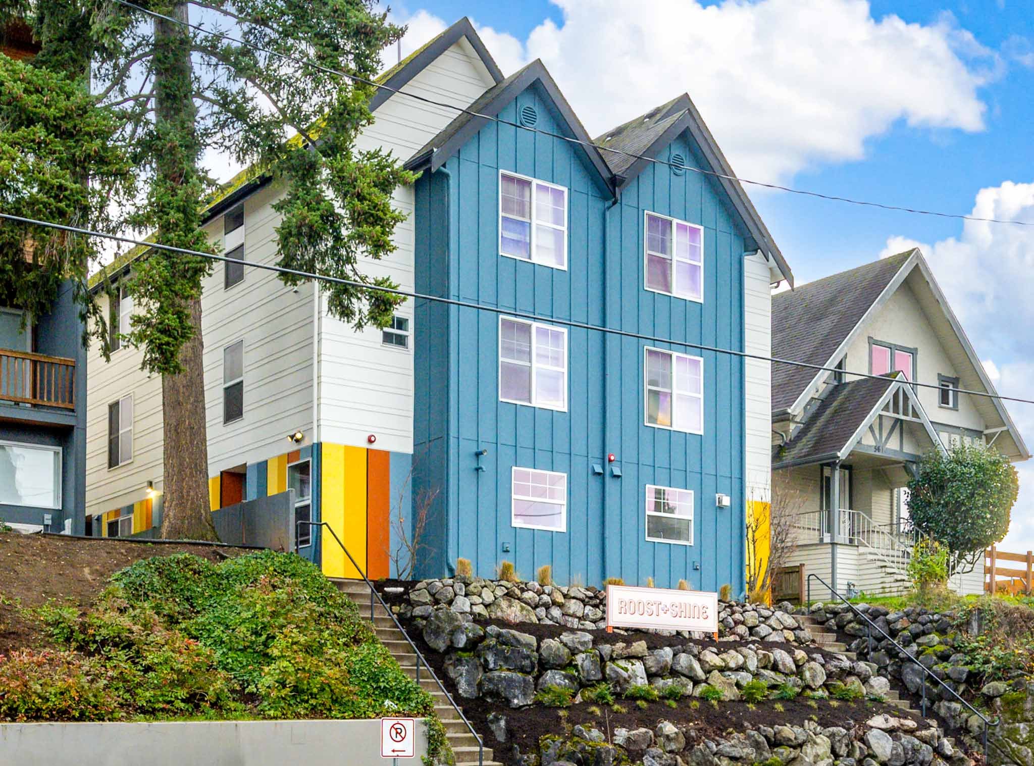 Colorful modern house with blue, white, and yellow panels, situated on a hillside with steps leading to the entrance and a sign that reads 'ROOST SHINE.' The house is next to a large tree, and the sky is partly cloudy.
