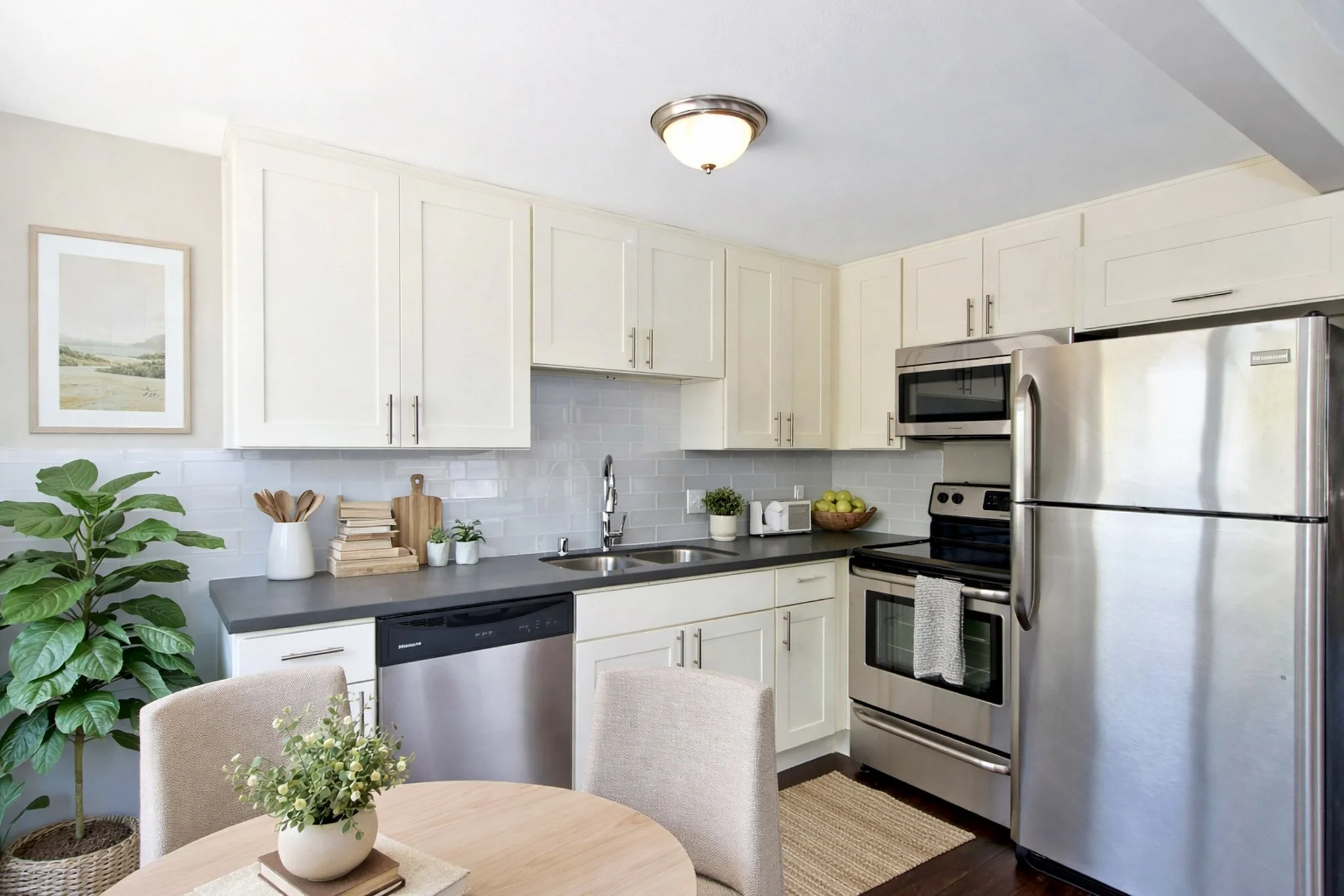 A modern kitchen with white cabinets, stainless steel appliances, a wooden dining table with beige chairs, a potted plant, and decorative items on the countertop.