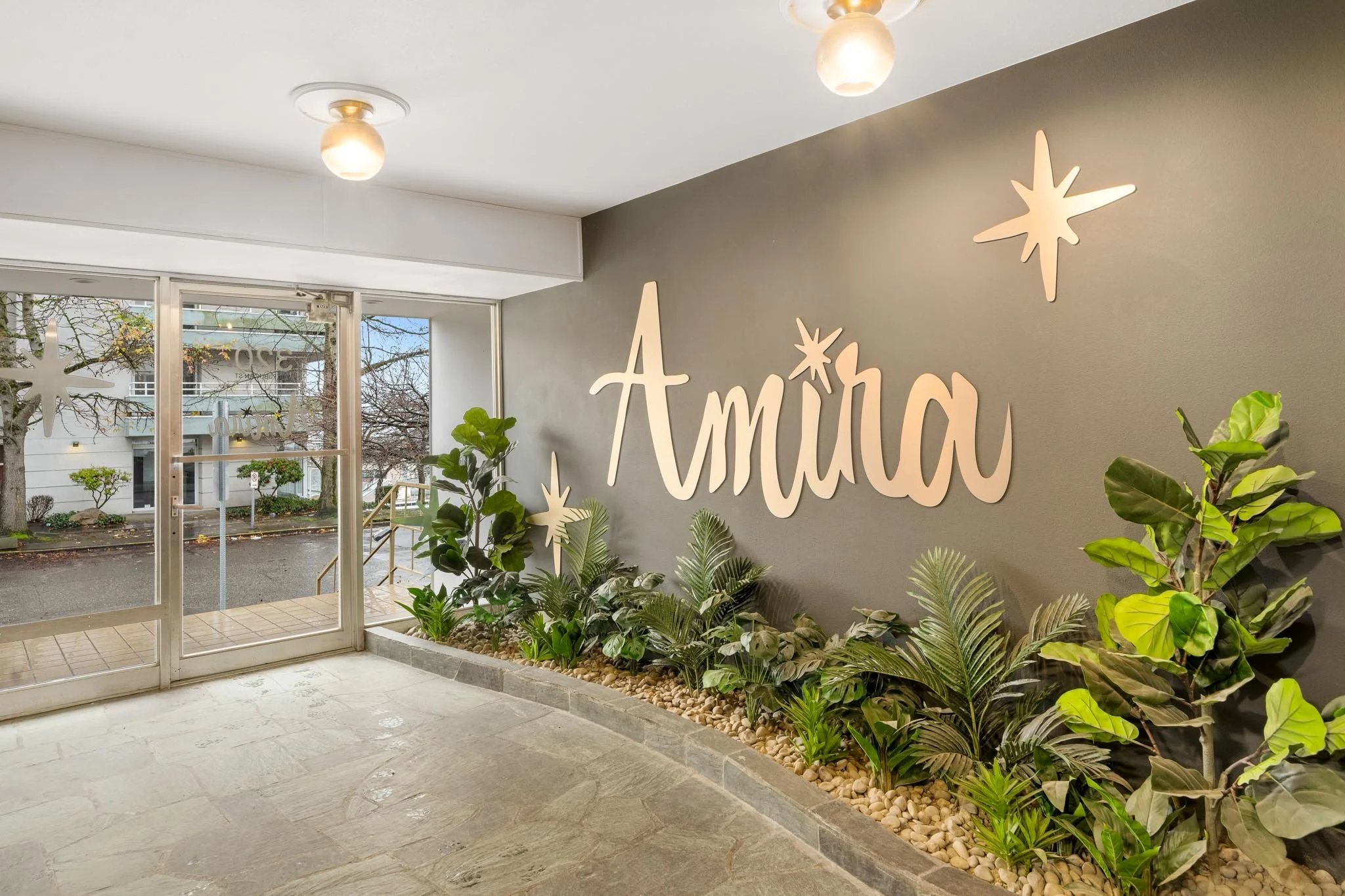 Interior of a building with a glass door entrance, decorated with white star-shaped ornaments and the name 'Amoa' on the wall. There are plants along the wall near the entrance and a view of trees and buildings outside.