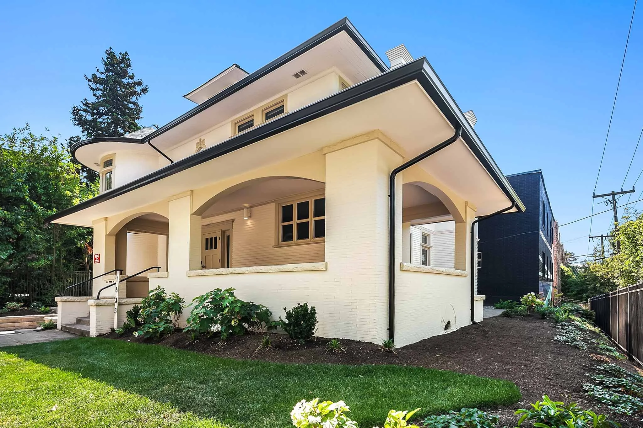 A two-story house with a light beige exterior, large front porch, and black roof trim. The front yard has a green lawn, bushes, and small plants, with a pathway and steps leading to the front door.