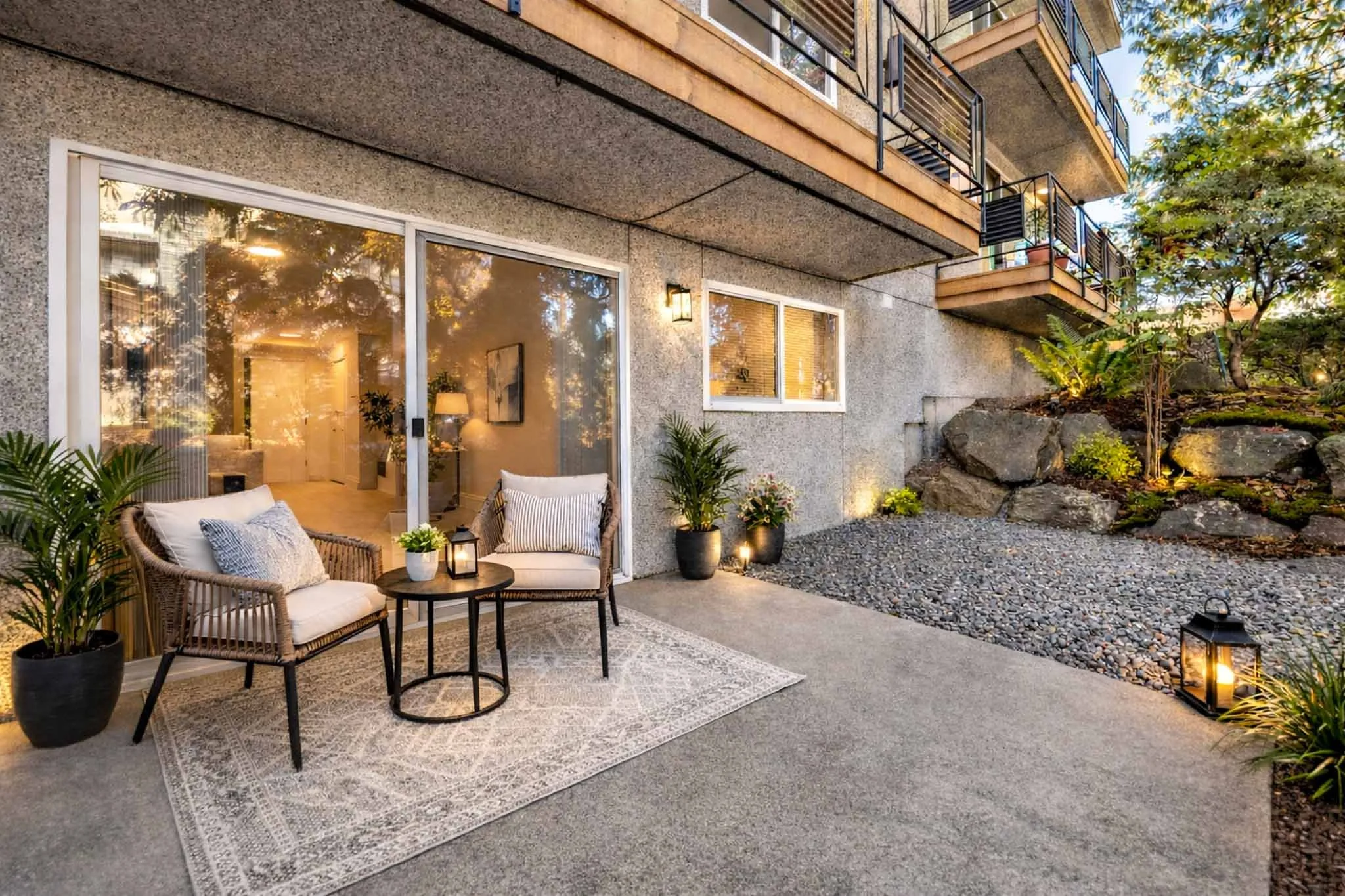An outdoor patio area of an apartment with cozy seating, a rug, potted plants, and warm lighting, adjacent to large glass sliding doors leading inside. There is a garden with rocks and trees surrounding the space.