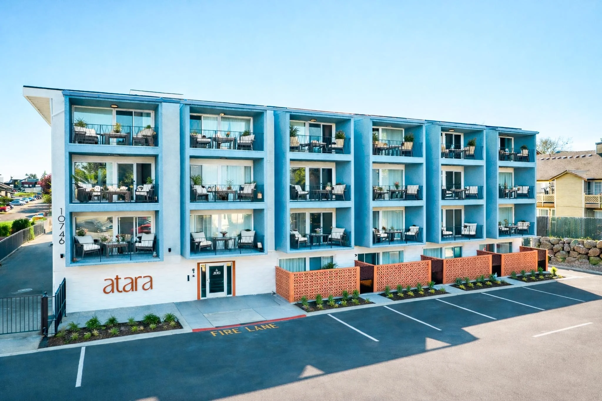Three-story building with balconies, light blue exterior, and patio furniture on each balcony. Parking lot in front with designated fire lane, and some greenery and rocks along the sidewalk.