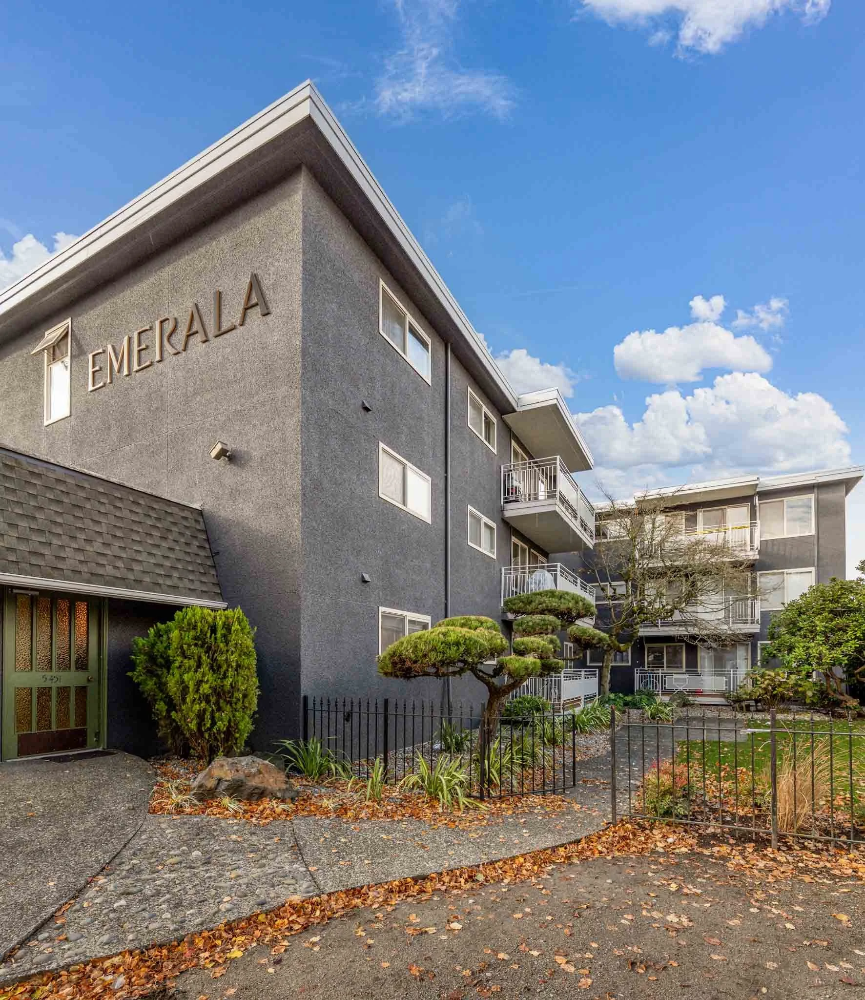 View of a gray multi-story apartment building labeled 'EMERALD,' with balconies, surrounded by a garden with trimmed trees, bushes, and fallen leaves, under a partly cloudy blue sky.