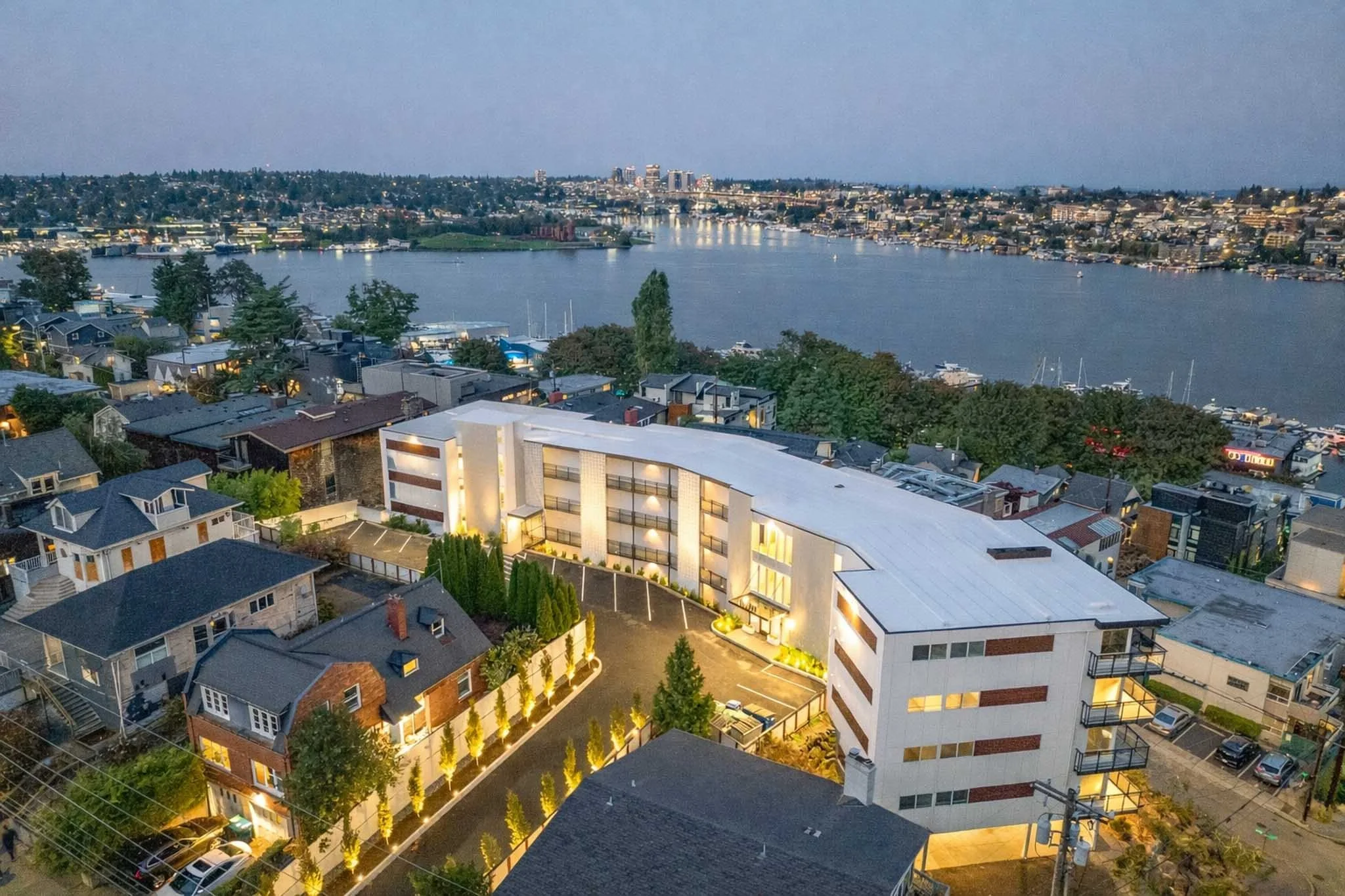 Cityscape at dusk featuring a river with boats, surrounding residential and commercial buildings, and a modern white building with lit windows in the foreground.
