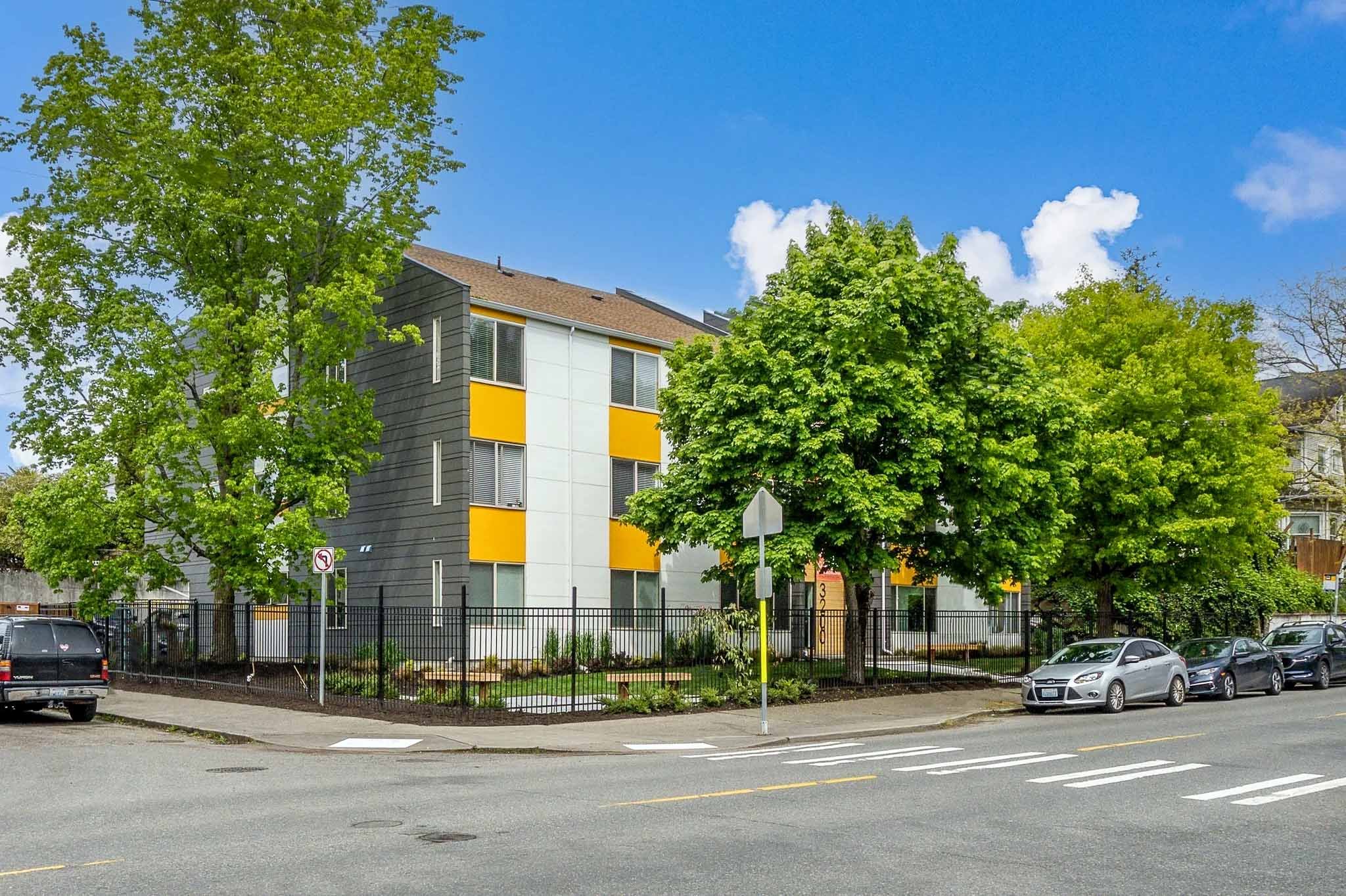 A modern apartment building with yellow and white accents, surrounded by green trees, under a bright blue sky with some white clouds. There is a parking lot with several cars in front, and a street in the foreground.