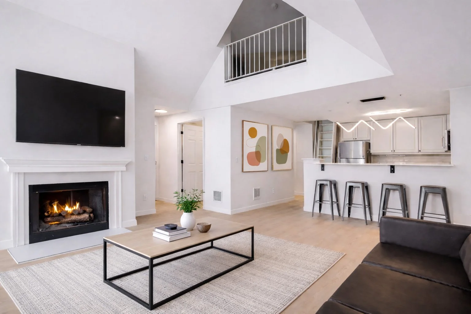 Modern living room with white walls, a fireplace, a mounted flat-screen TV, a coffee table with decorative items, a painting on the wall, a black sofa, and a kitchen area with barstools and abstract art