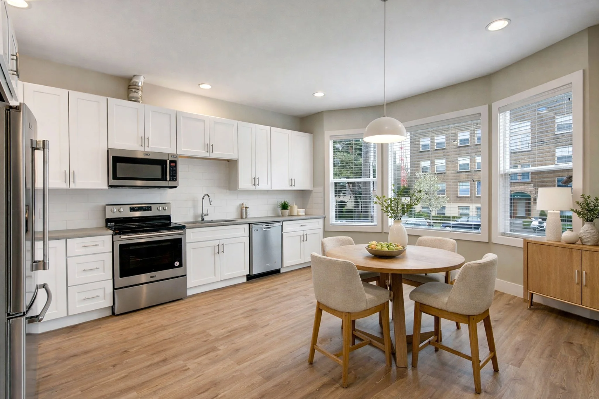 A modern kitchen with white cabinets, stainless steel appliances, and large windows letting in natural light. There's a round wooden dining table with four beige upholstered chairs, a white vase with greenery, and a bowl of apples on the table.