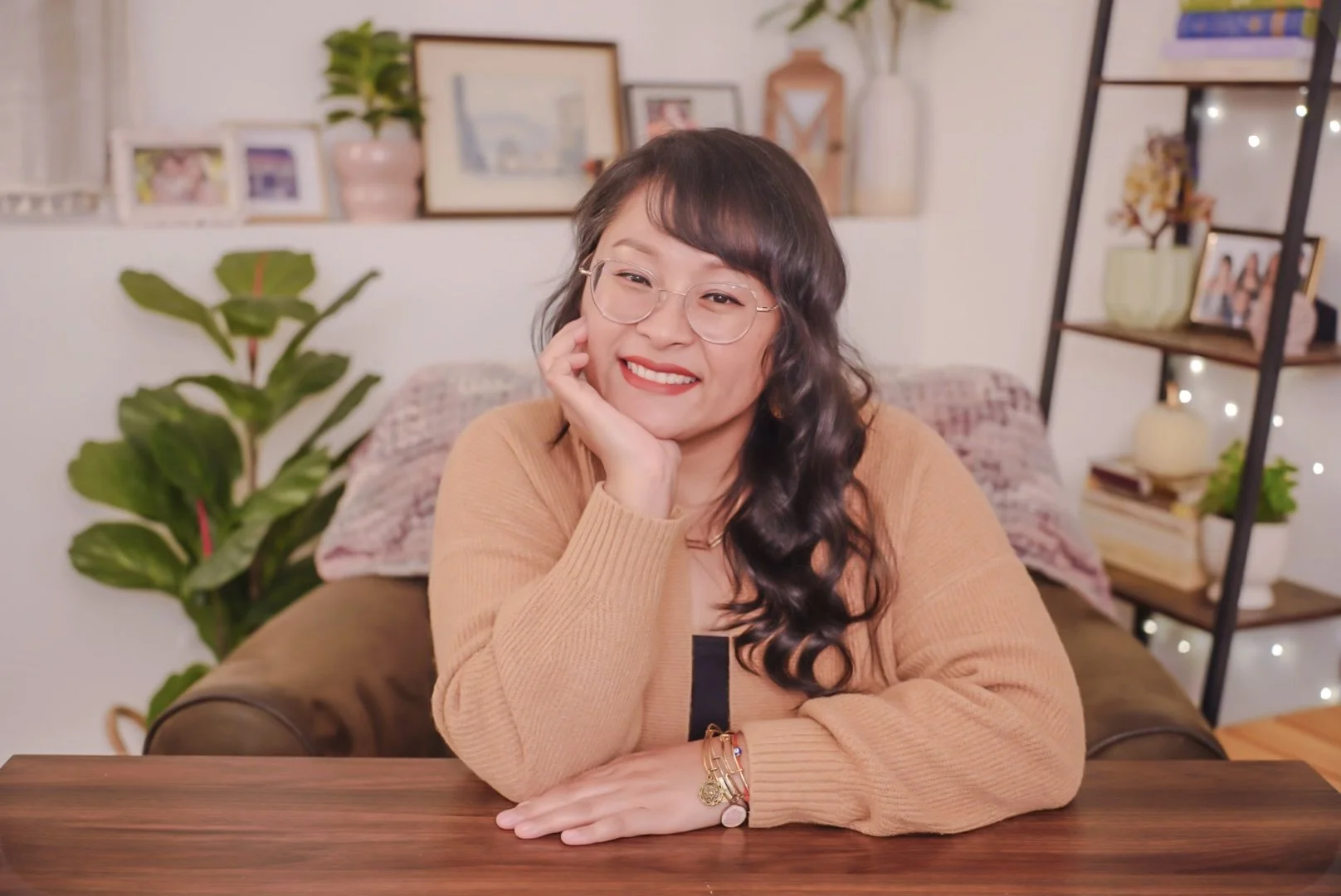 A woman with dark hair and glasses sitting at a table, smiling and resting her face on her hand in a cozy, decorated living room.