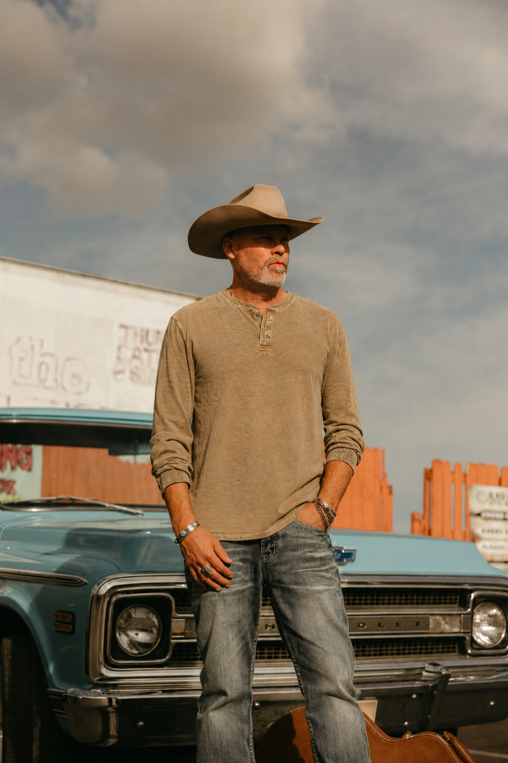 A man wearing a cowboy hat and casual clothing stands in front of a vintage car, with a partly cloudy sky overhead.