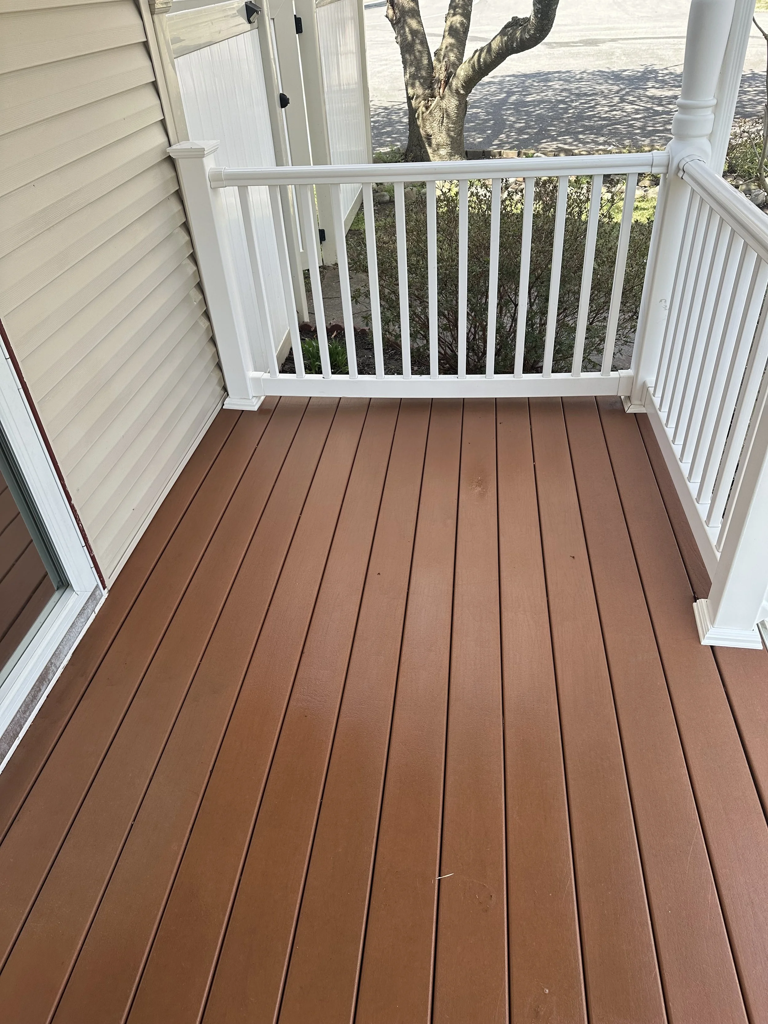 Empty balcony with wood flooring, white railing, beige siding, and a tree outside.