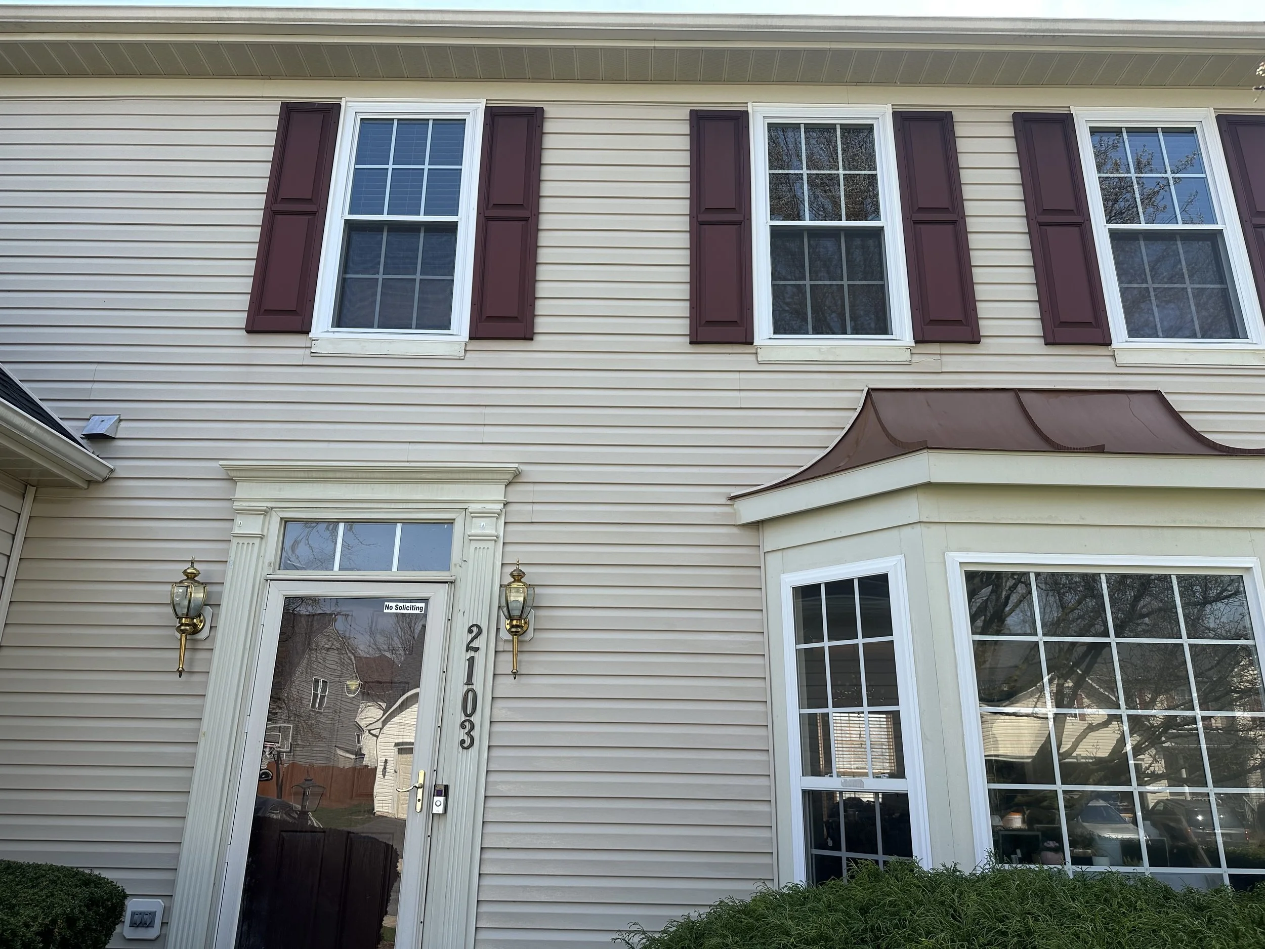 Front view of a beige two-story house with purple shutters, a bay window, and a glass front door with the house number 2103 and a 'No Soliciting' sign.