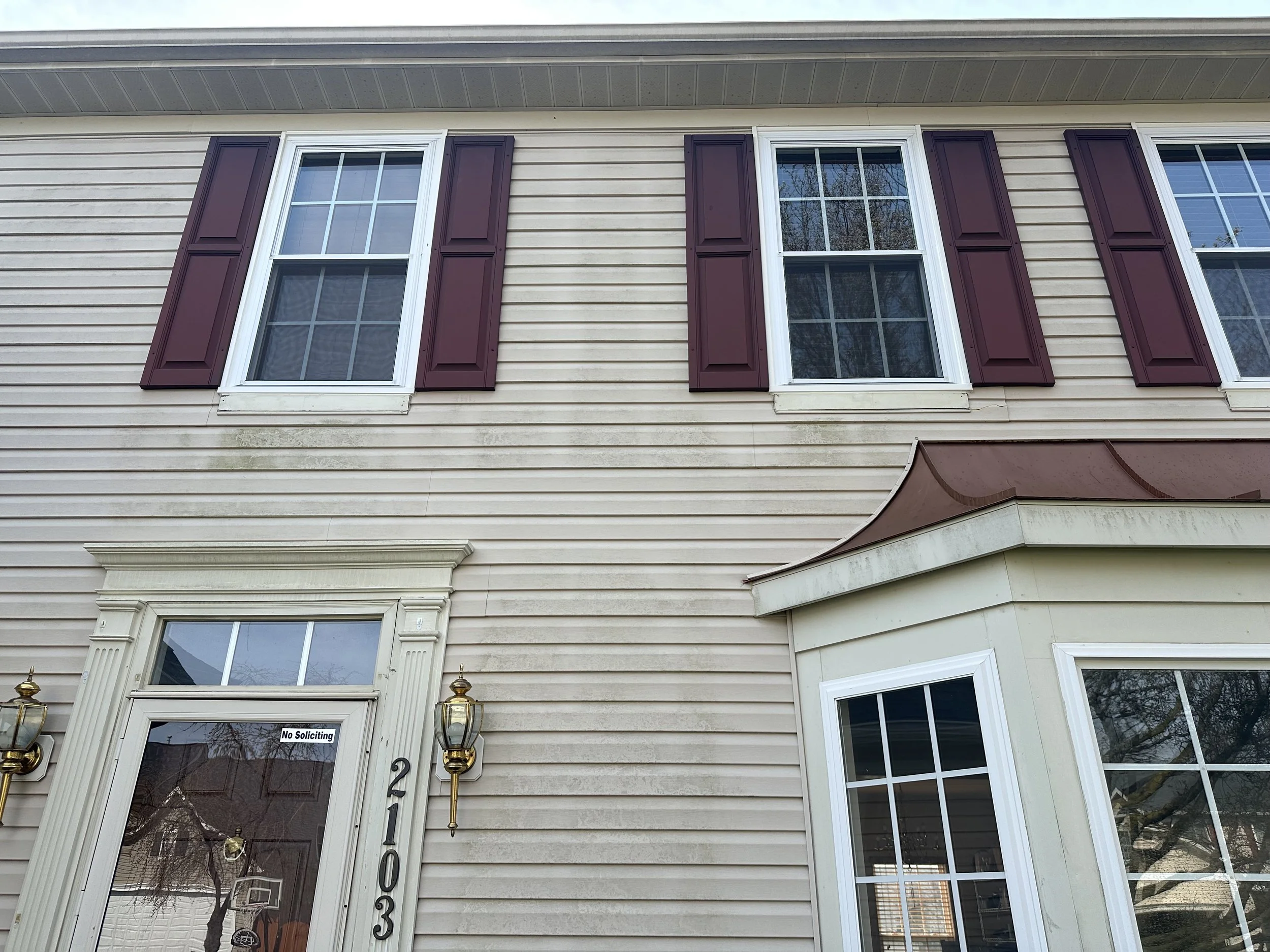 Front exterior of a beige house with white trim, three windows with maroon shutters, and a double front door with a sign that says 'No Soliciting'.