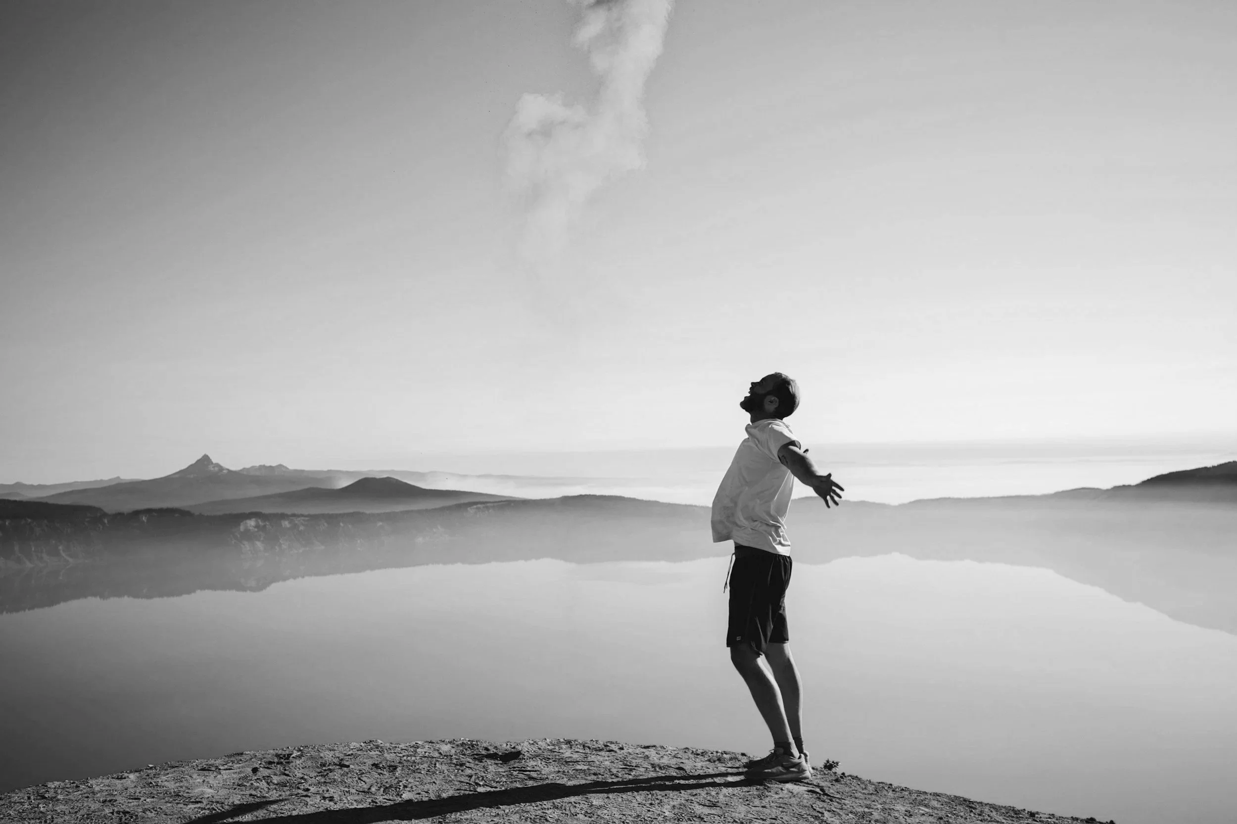A person standing on a rocky ledge with arms stretched out, facing a calm body of water with mountains in the background and a cloudy sky, captured in black and white.