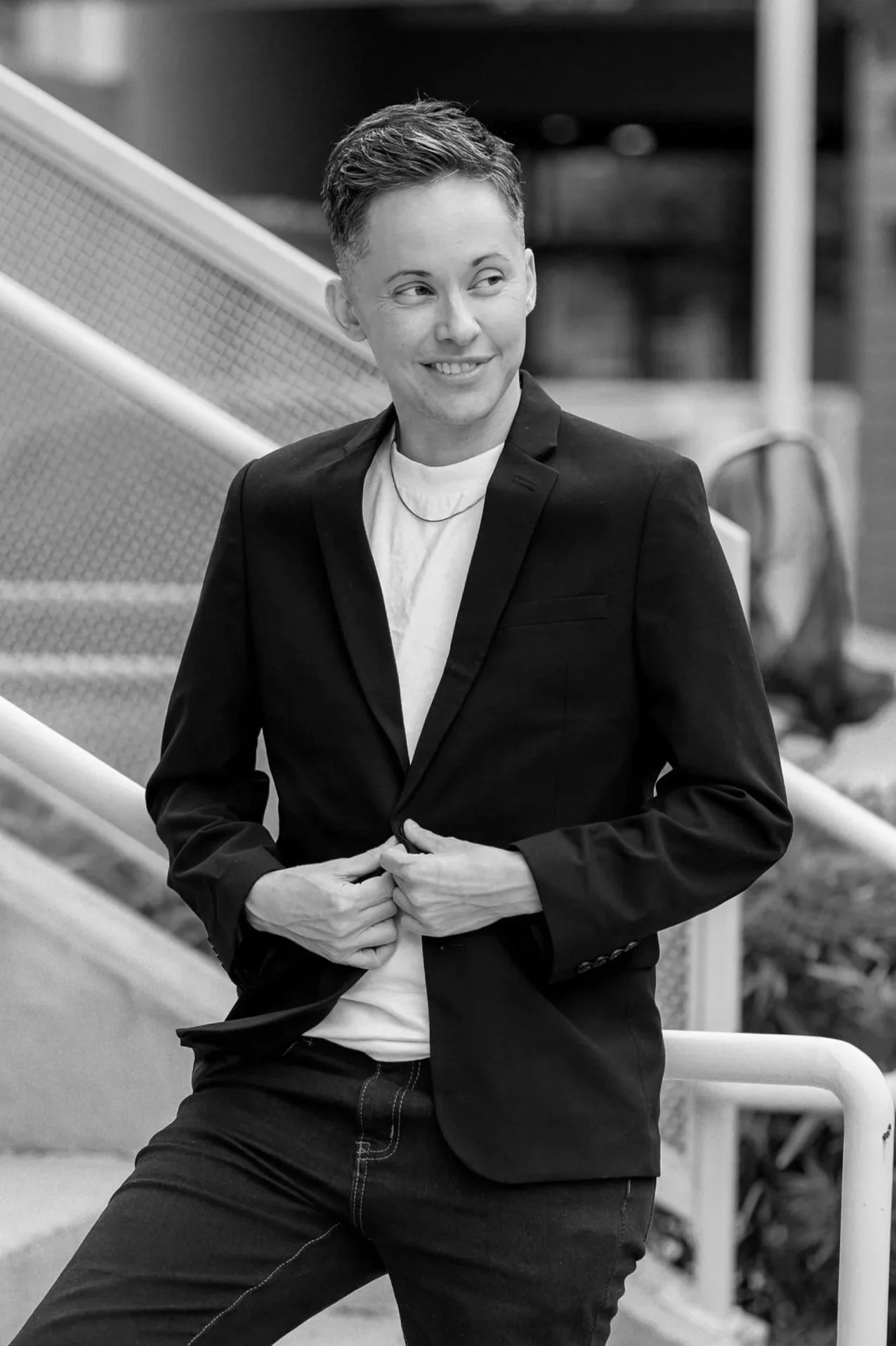 A woman with short hair, wearing a blazer and a white t-shirt, smiling and sitting outdoors near a stairway, in a black-and-white photo.