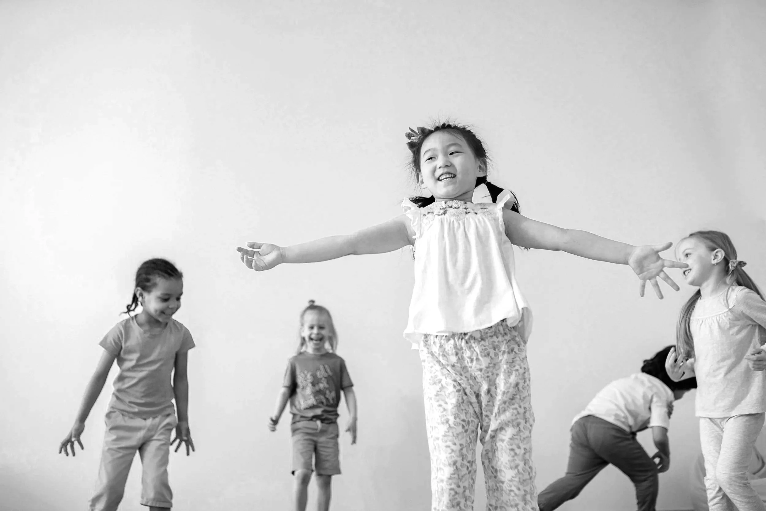 Black and white photo of five young children playing and laughing indoors, with a plain wall background.