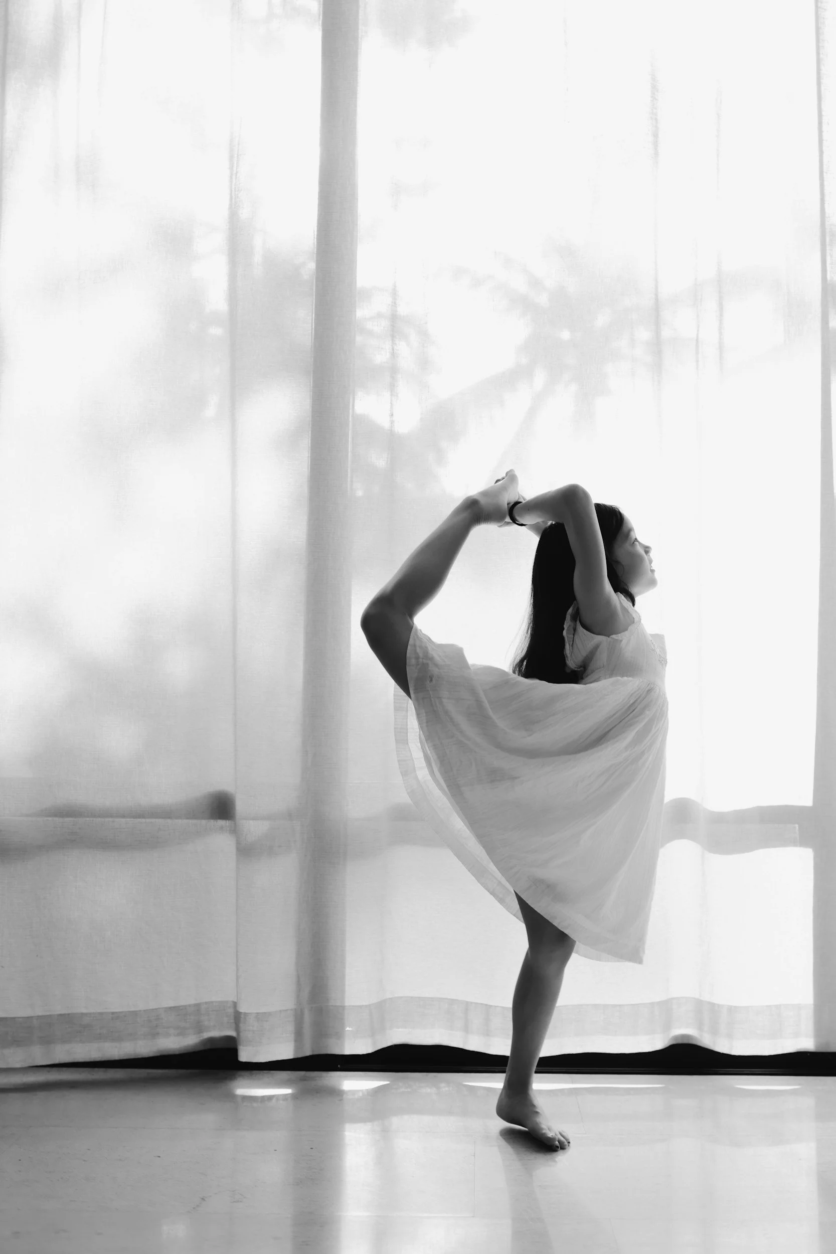 A woman in a light dress is performing a dance pose indoors against a backdrop of sheer curtains with trees outside visible. She is balancing on one foot with the other leg bent behind her, holding her foot with both hands behind her head.