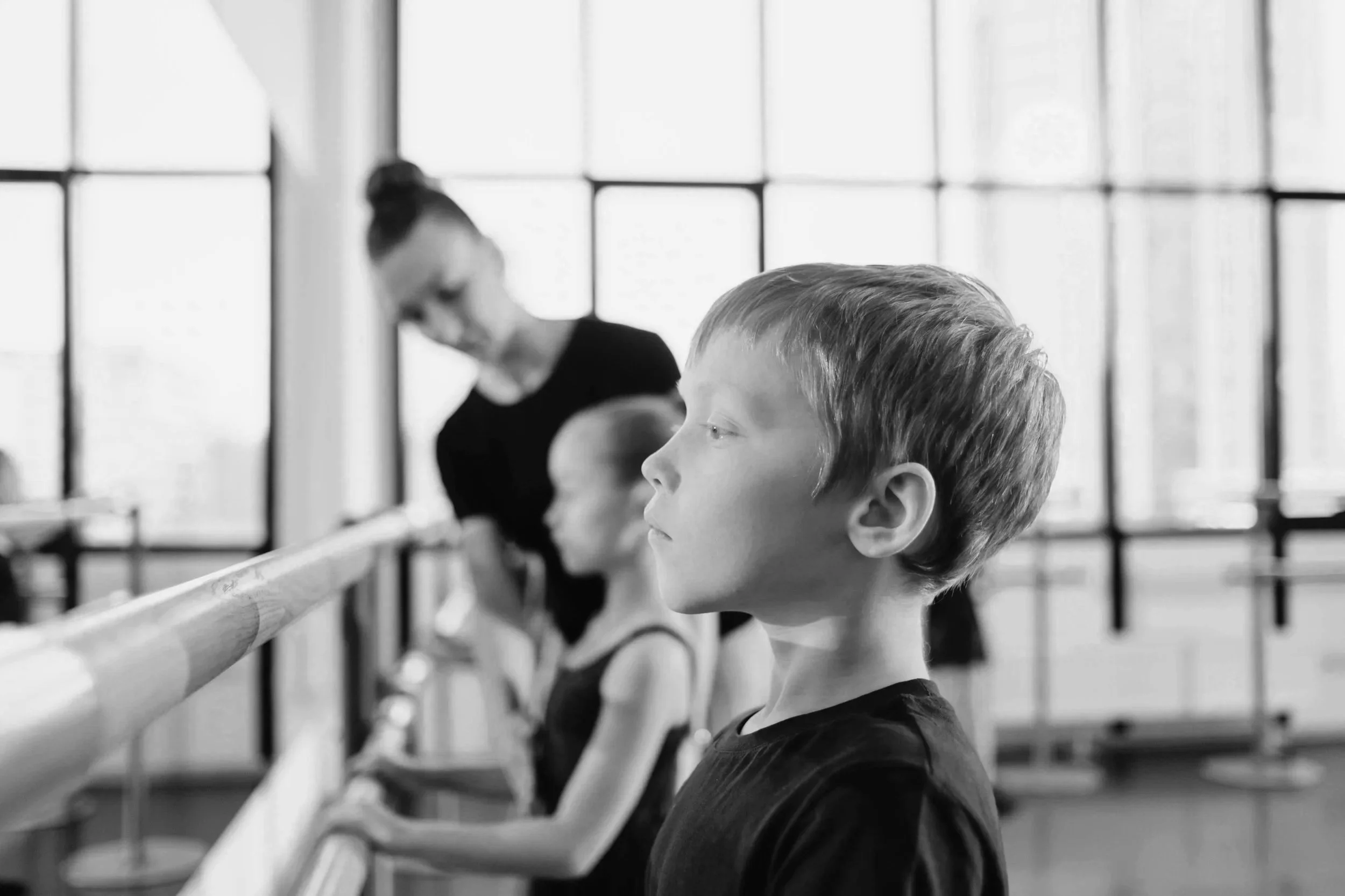 A boy with short hair in profile, standing at a ballet barre in a dance studio. Behind him, a girl and an adult woman are also engaged in dance practice, with large windows in the background.