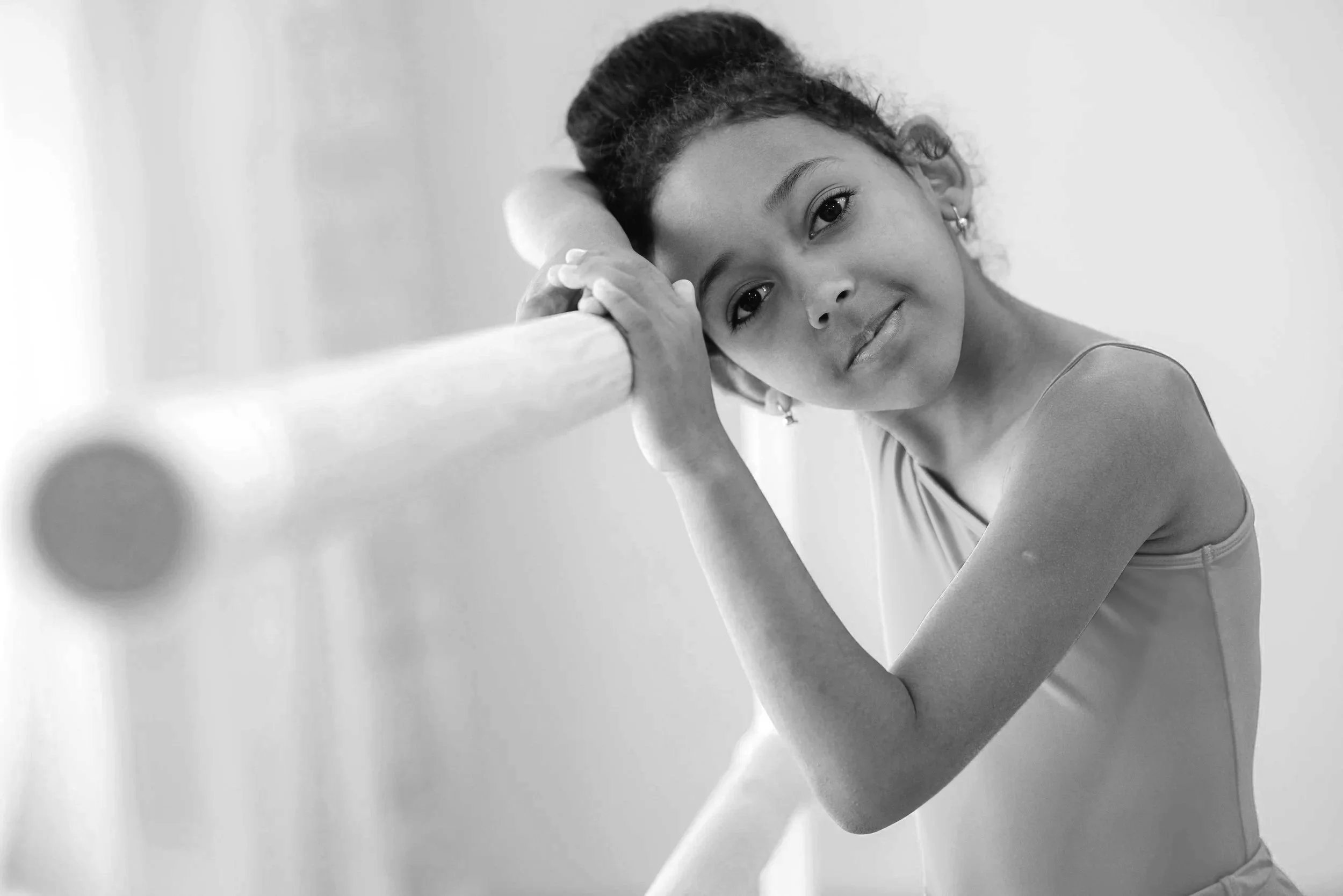 Young girl with braided hair leaning on a ballet barre, smiling softly, in a dance studio.