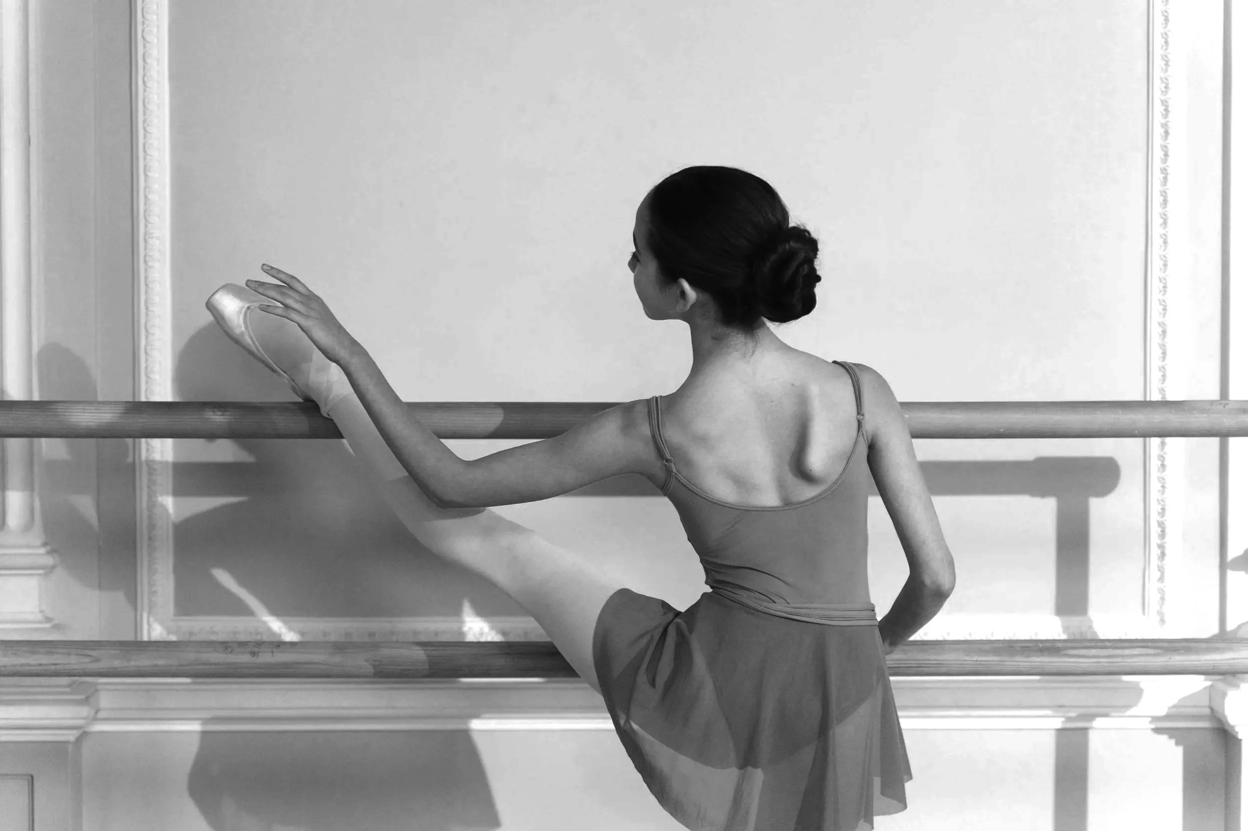 A ballerina stretching at a ballet barre, wearing a leotard and tutu, with her hair in a bun in a dance studio.