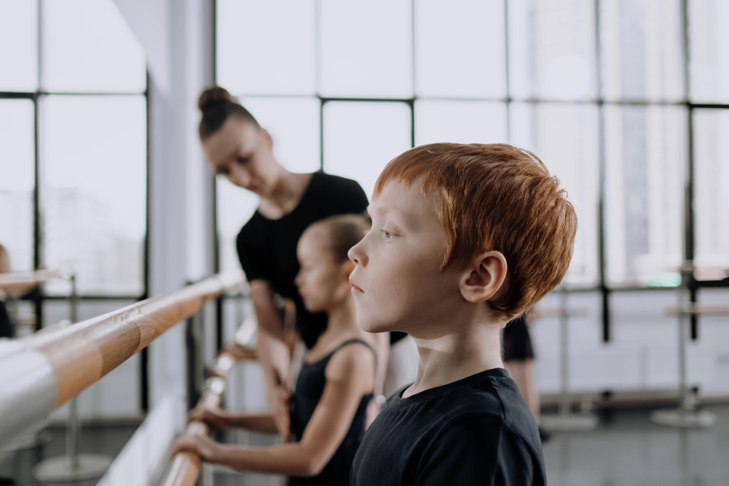 Young ballet students at the barre during a dance class, with a teacher assisting in the background.