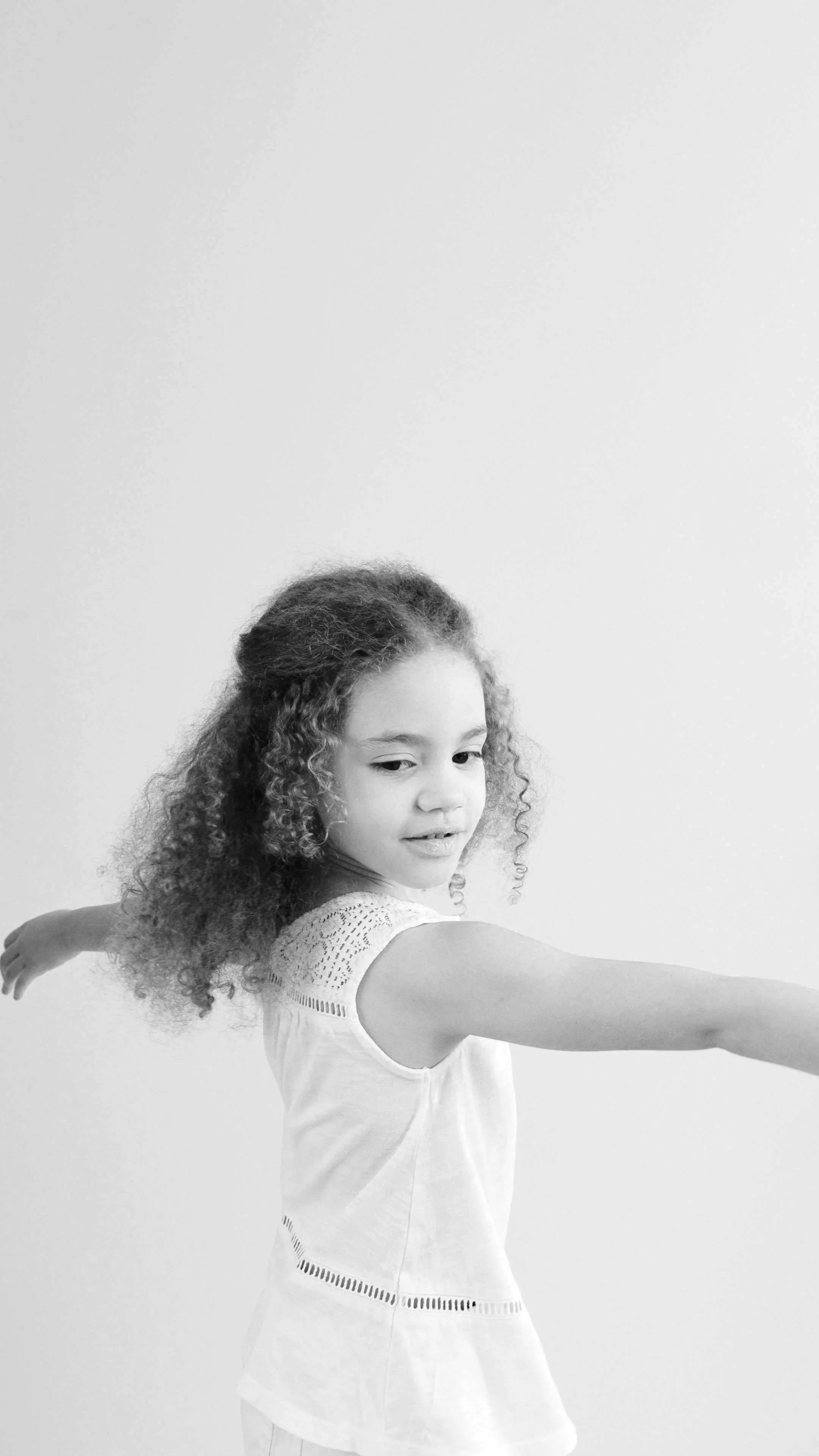 A young girl with curly hair in a white sleeveless top extending her arms to the sides.