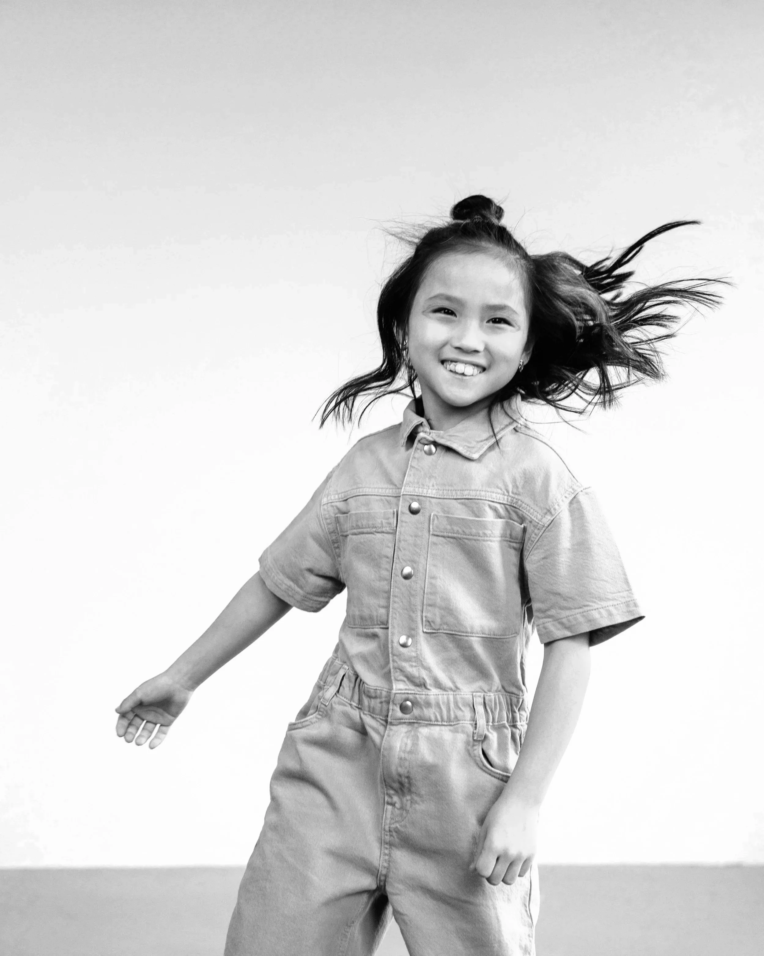 A young girl smiling and dancing, with her hair flowing, wearing a denim jumpsuit, against a plain background.