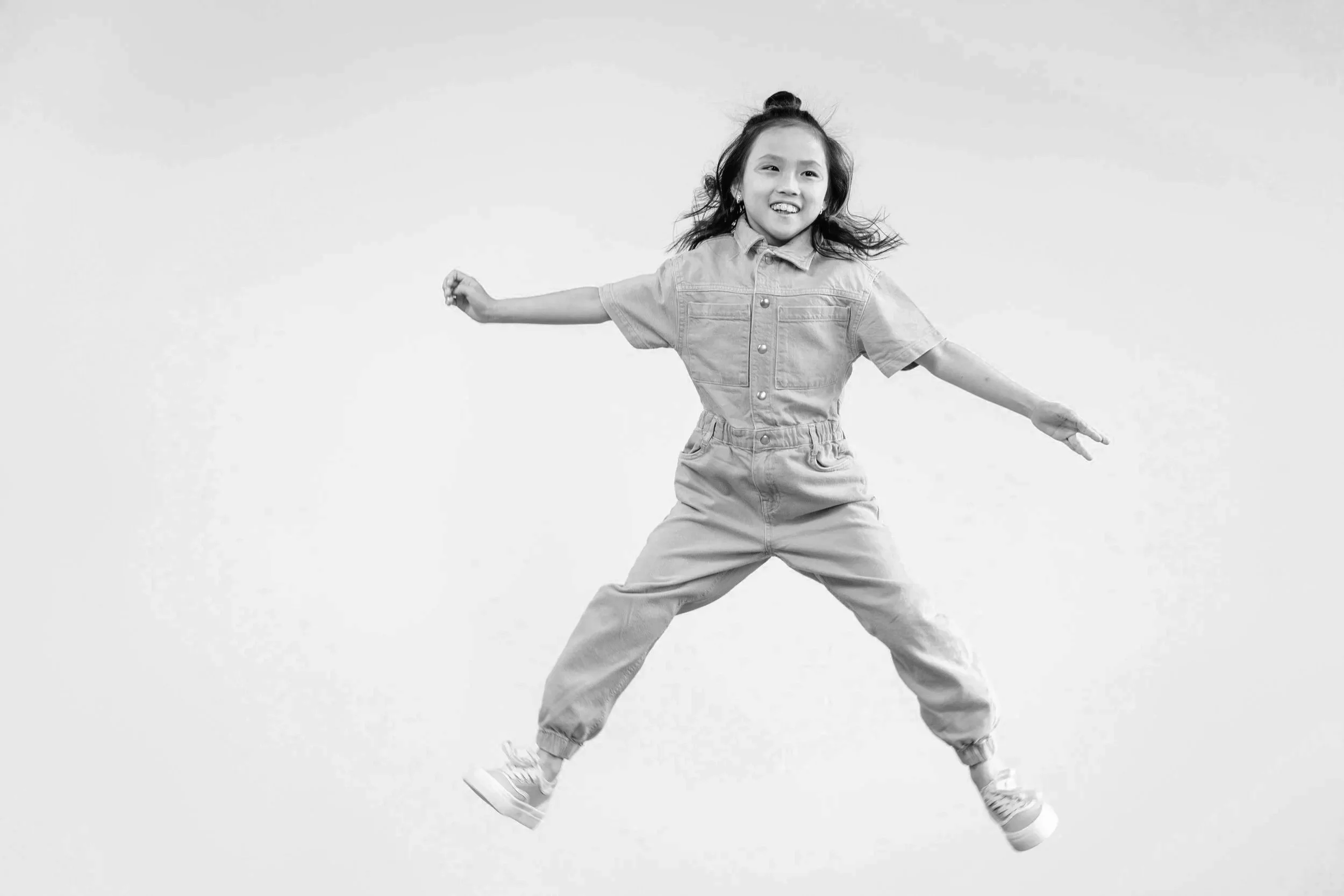 A young girl jumping in the air with her arms outstretched, wearing a jumpsuit and sneakers, smiling against a plain background.