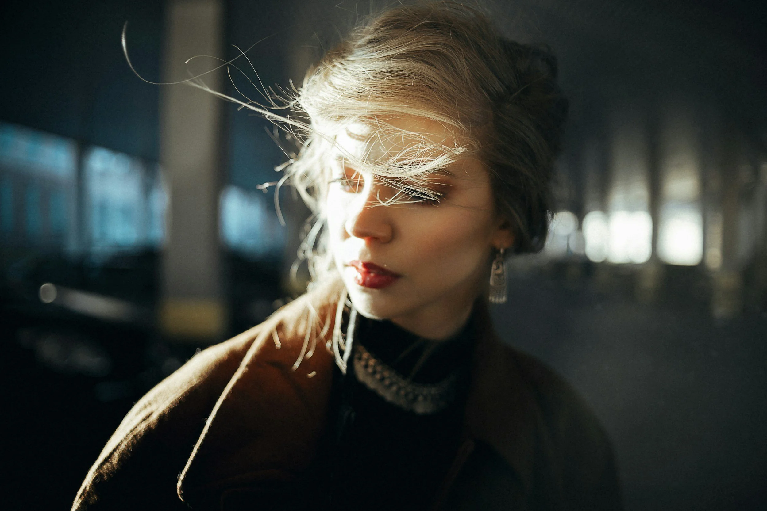 A woman with blonde hair in a ponytail and earrings, with closed eyes, illuminated by sunlight in a dark indoor setting.