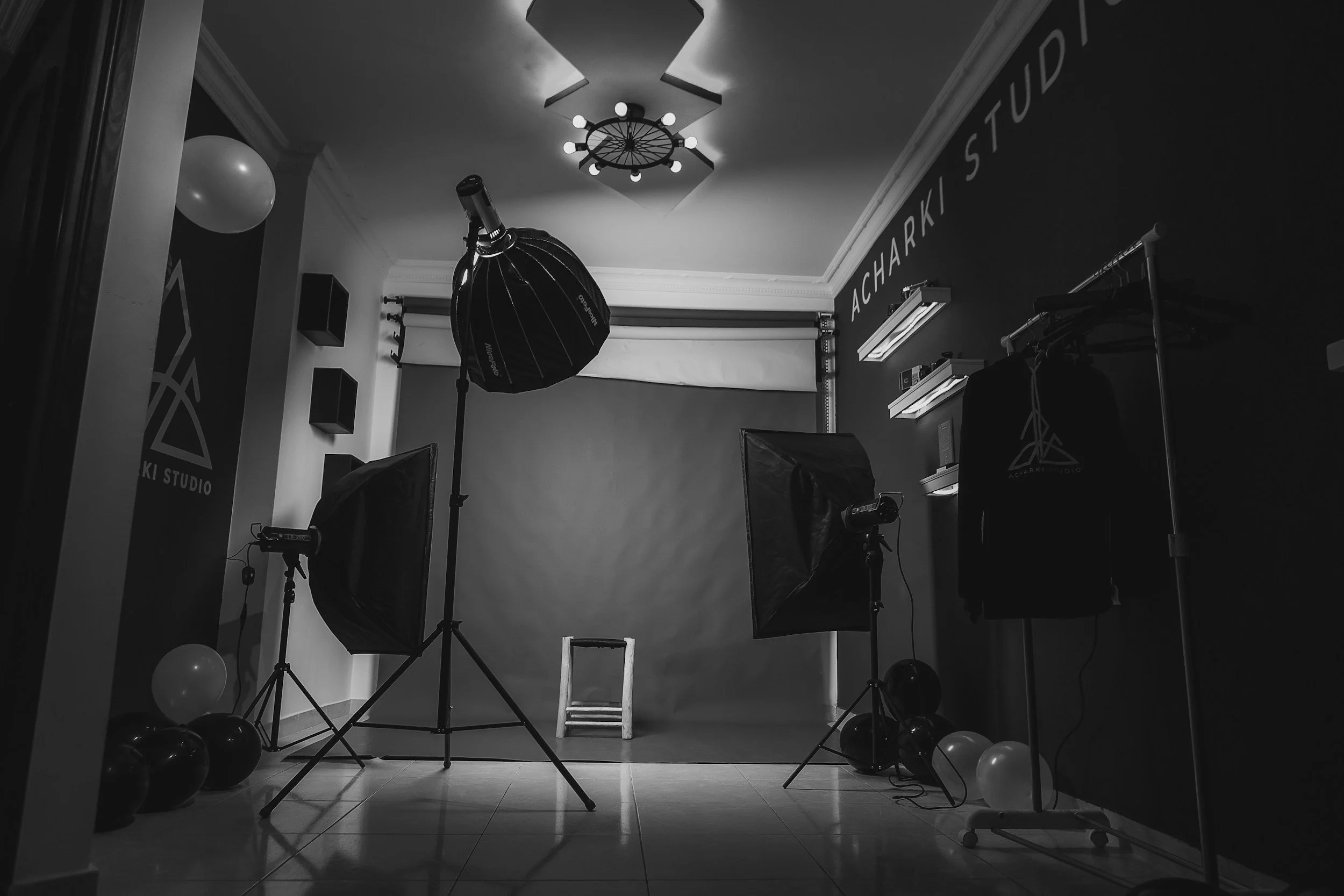 Photography studio setup with a gray backdrop, studio lighting, and balloons, inside a room with shelves, a hanging black hoodie, and the words 'ACHARSKI STUDIO' on the wall.