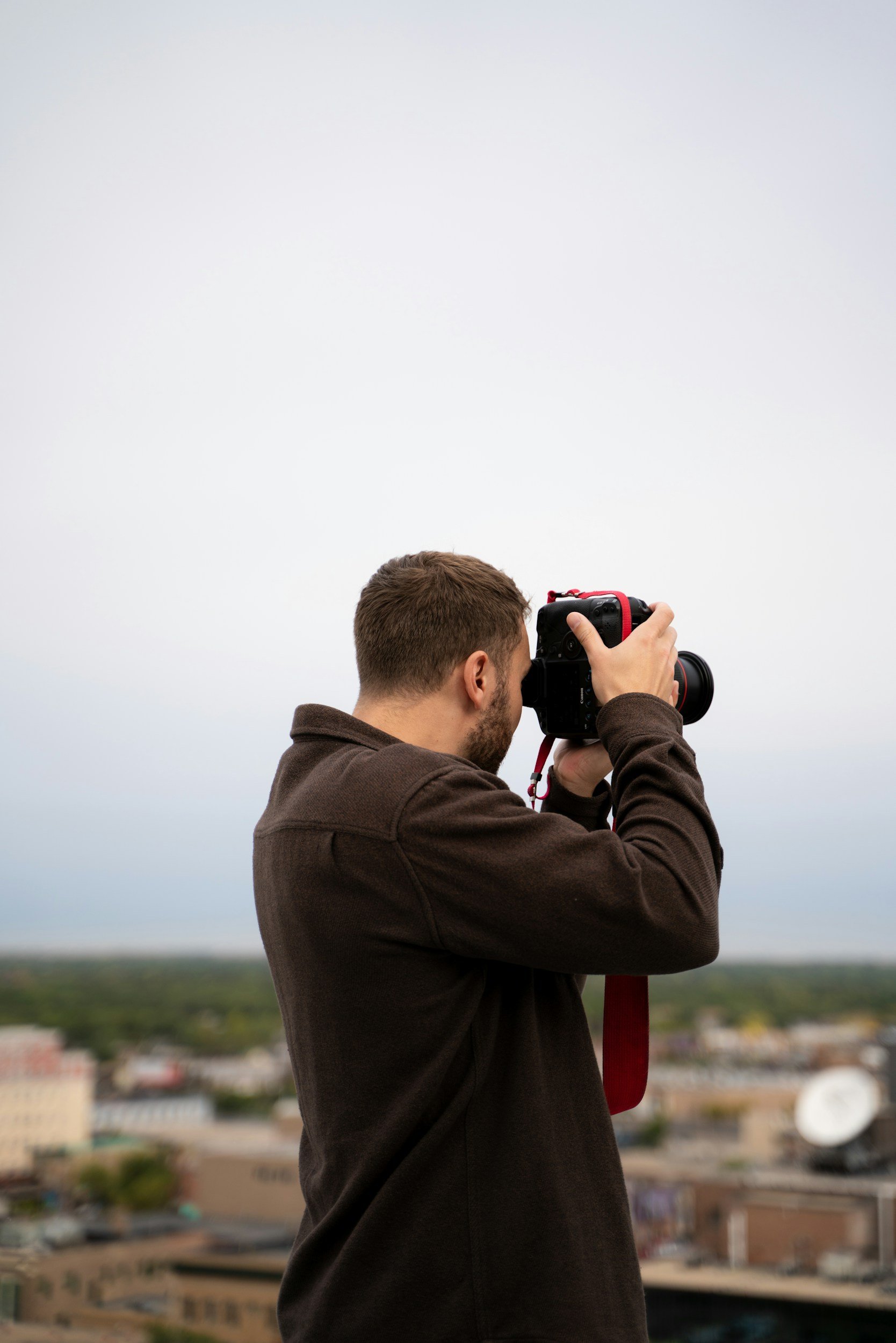 A man wearing a dark brown jacket is taking a photograph with a professional camera, facing a cityscape with buildings and a distant horizon under a cloudy sky.