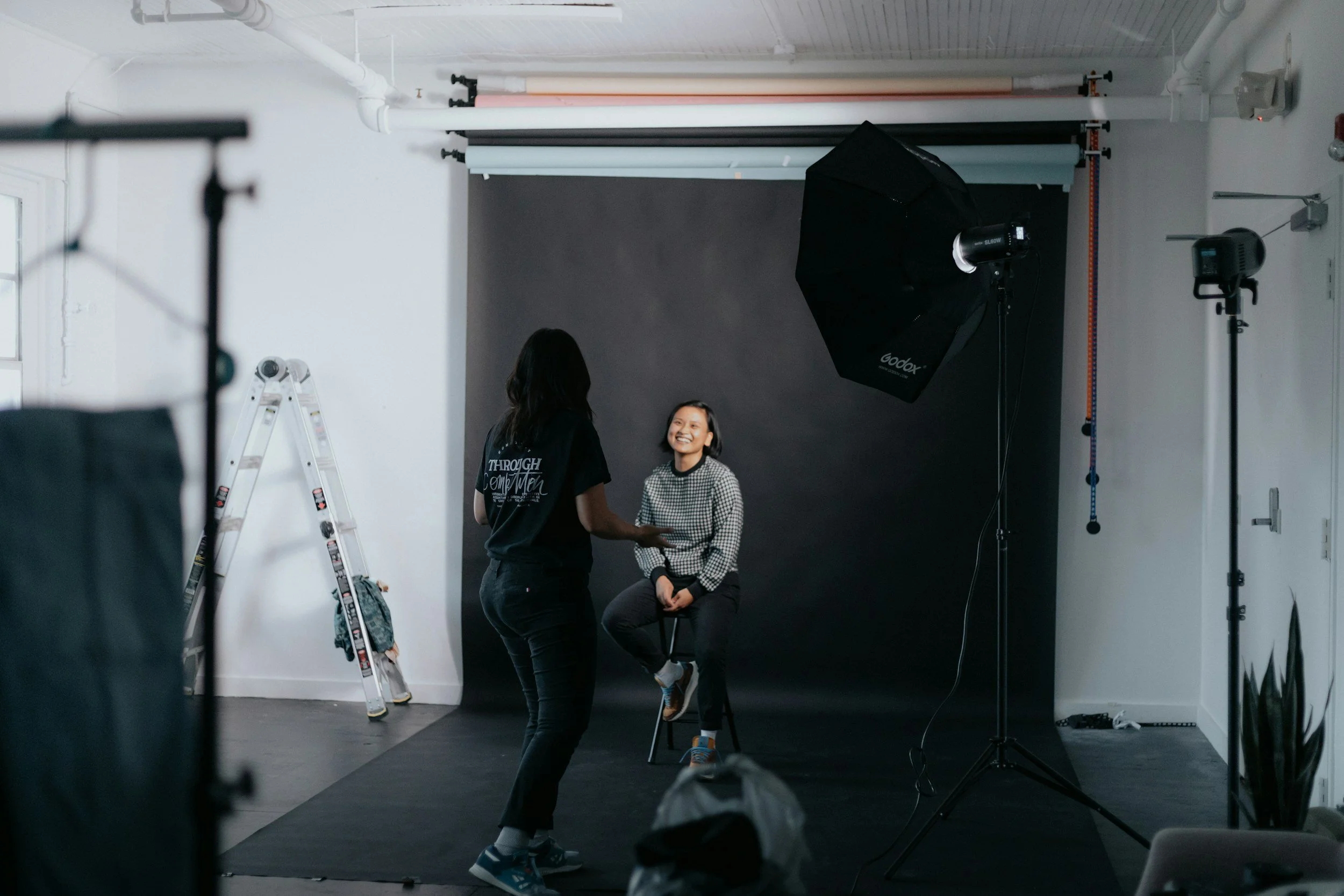A woman and a girl in a photography studio with a black backdrop, studio lights, and equipment.