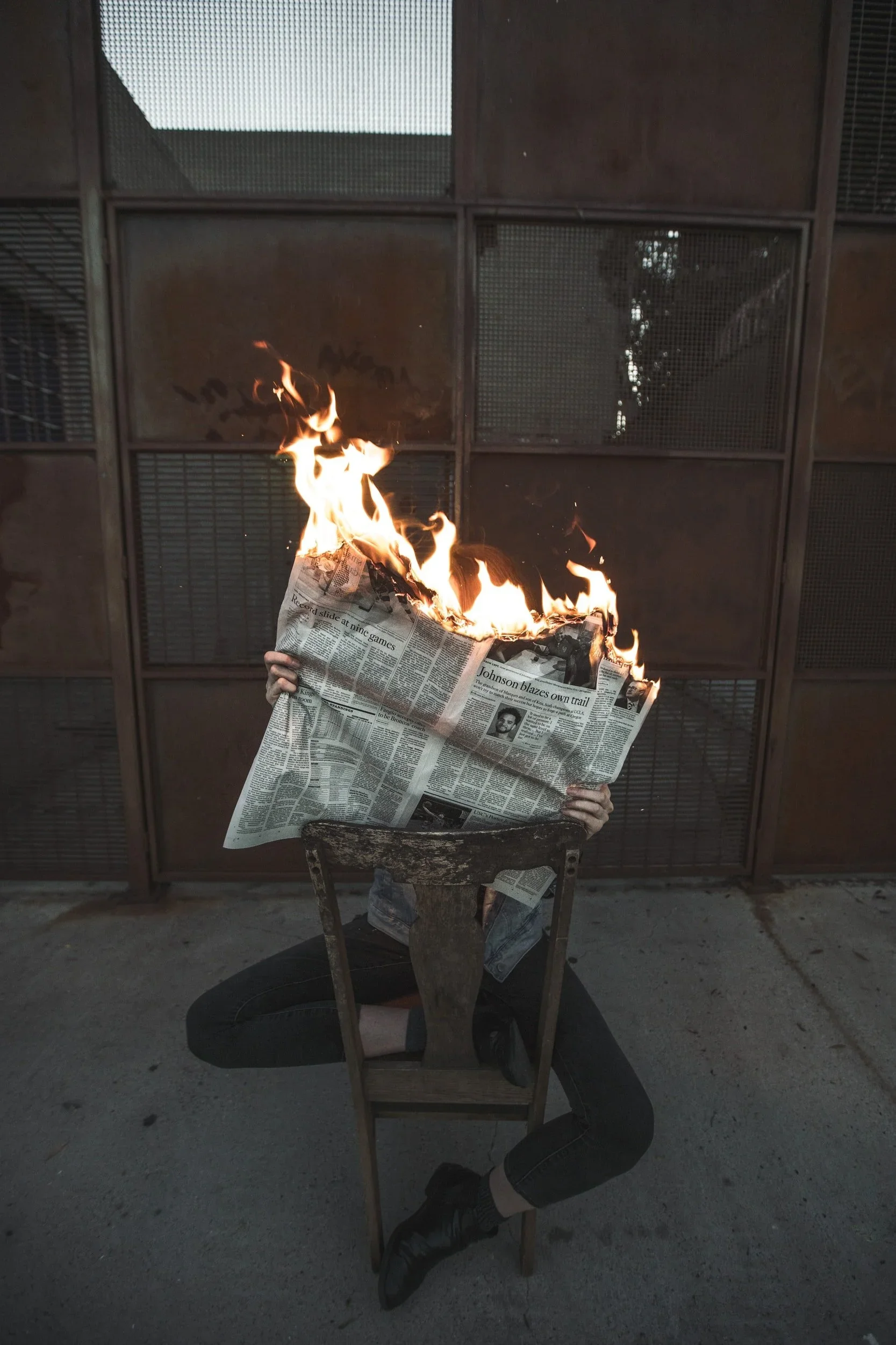 Person with newspaper on fire, sitting on a chair outdoors in front of a metal grid wall.
