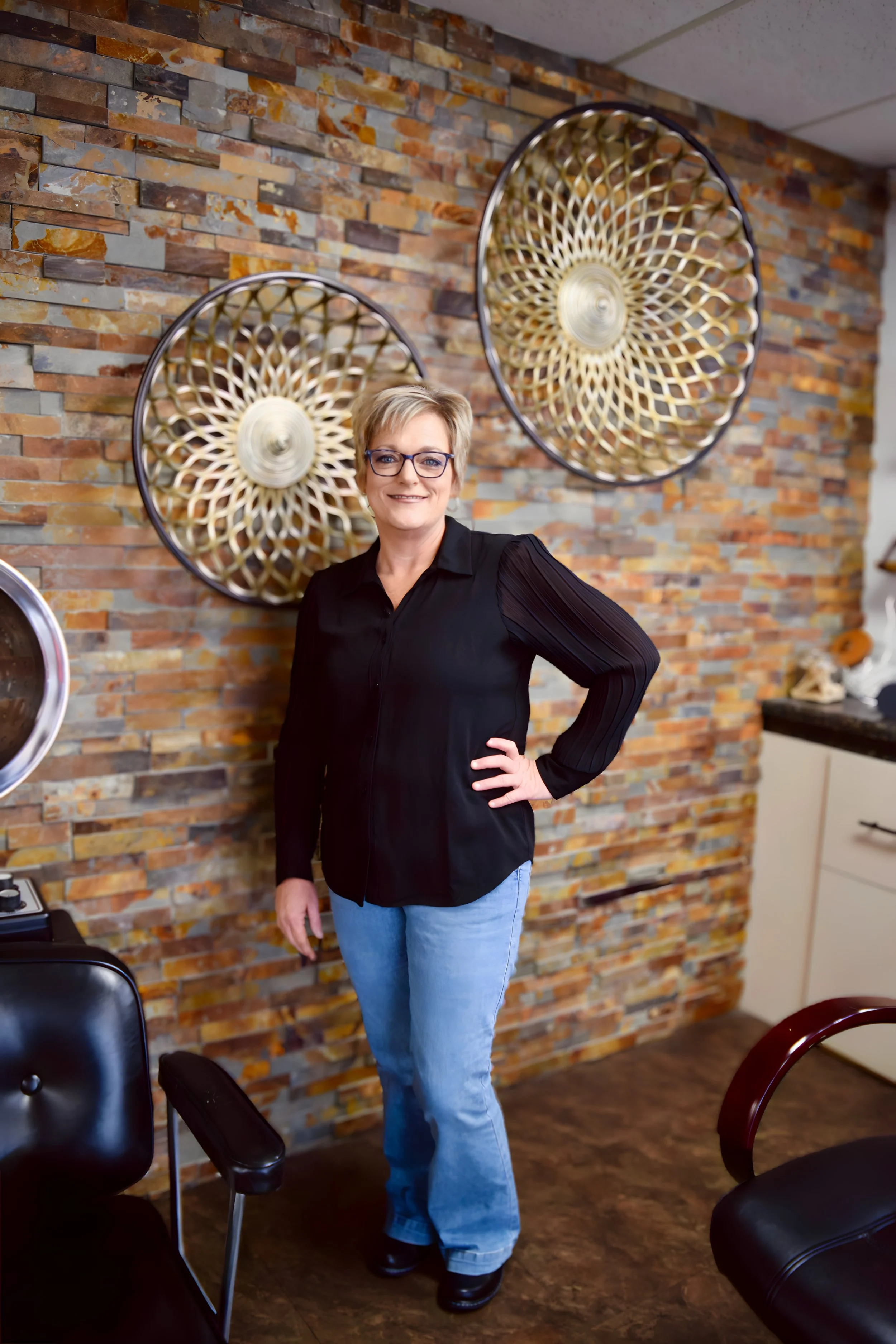 A woman with glasses, blonde hair, wearing a black shirt and light blue jeans, standing indoors with a brick wall behind her, decorated with two large circular woven wall hangings.