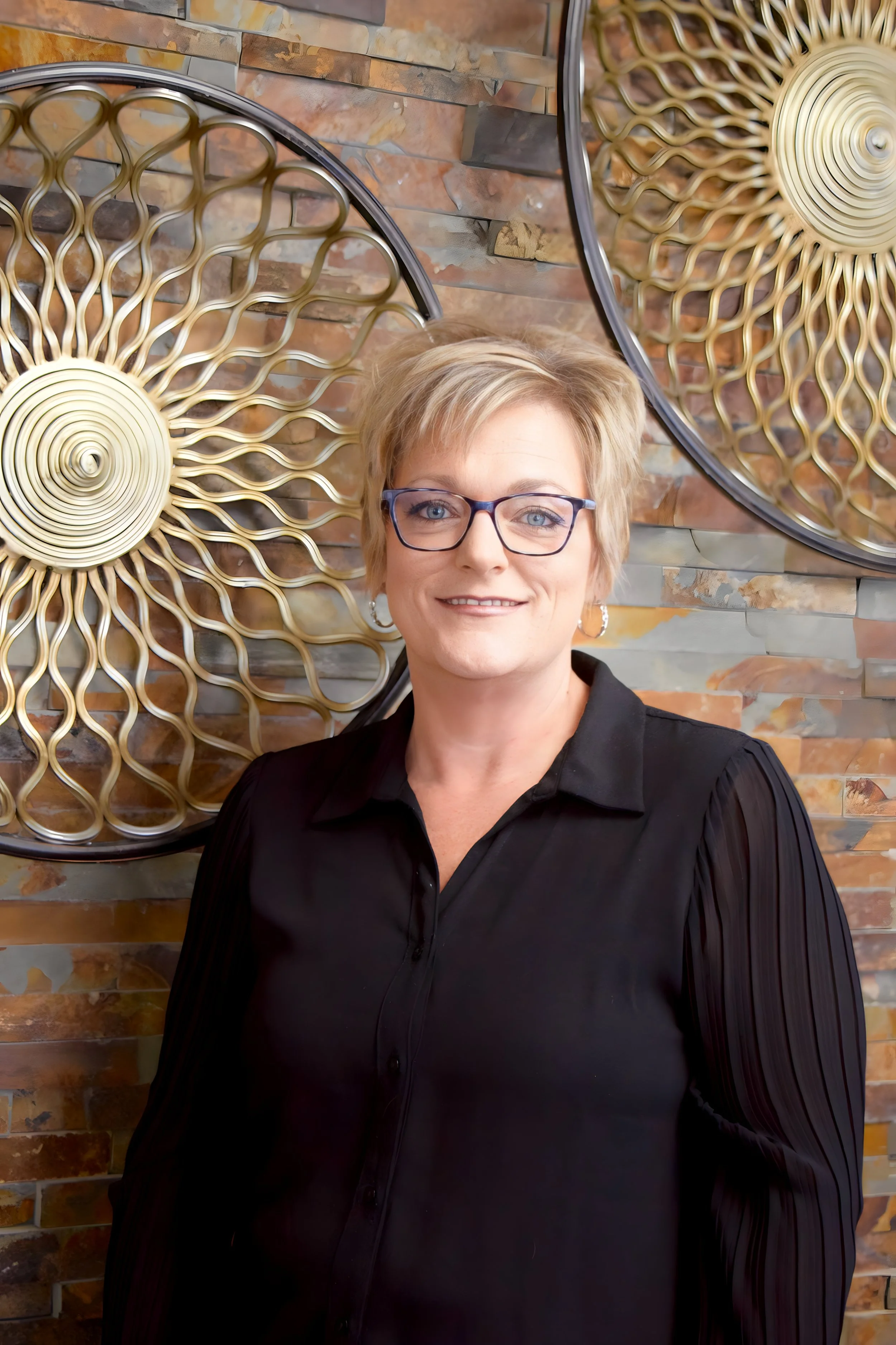 A woman with short blonde hair and glasses smiling in front of a decorative brick wall with gold-colored circular metal art pieces.