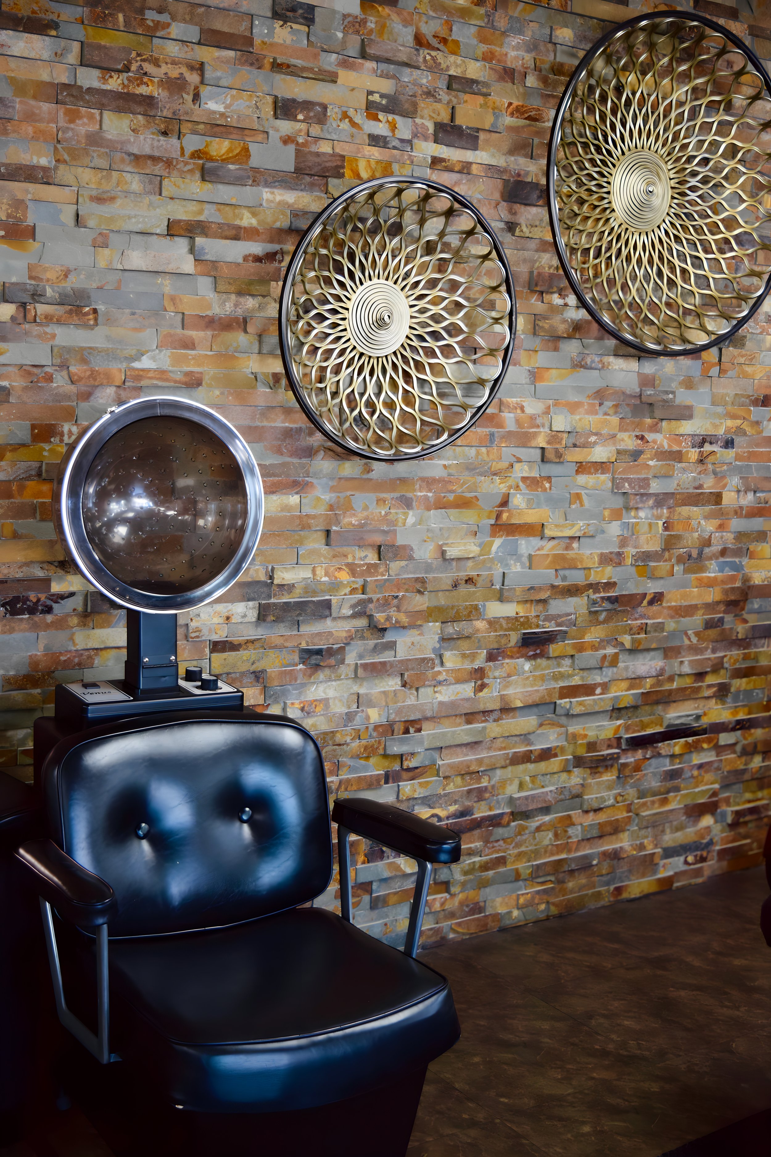 A vintage black salon chair with a metal frame next to a hair dryer on a stand. The setting has a brick wall with two decorative gold circular wall art pieces with intricate designs.