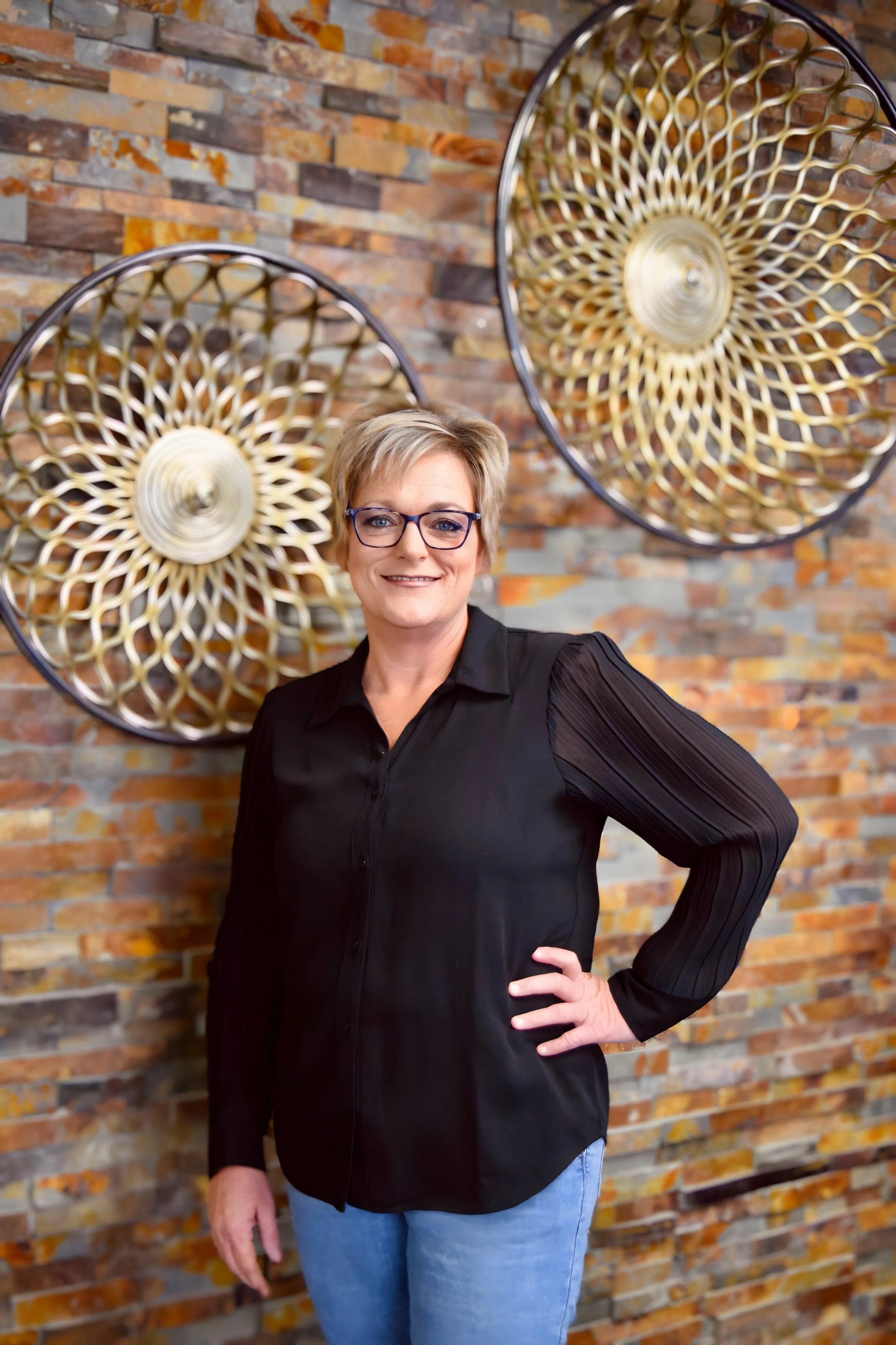 A woman with short blonde hair, wearing glasses, a black blouse with pleated sleeves, and blue jeans, standing in front of a brick wall with large decorative circular wall art.