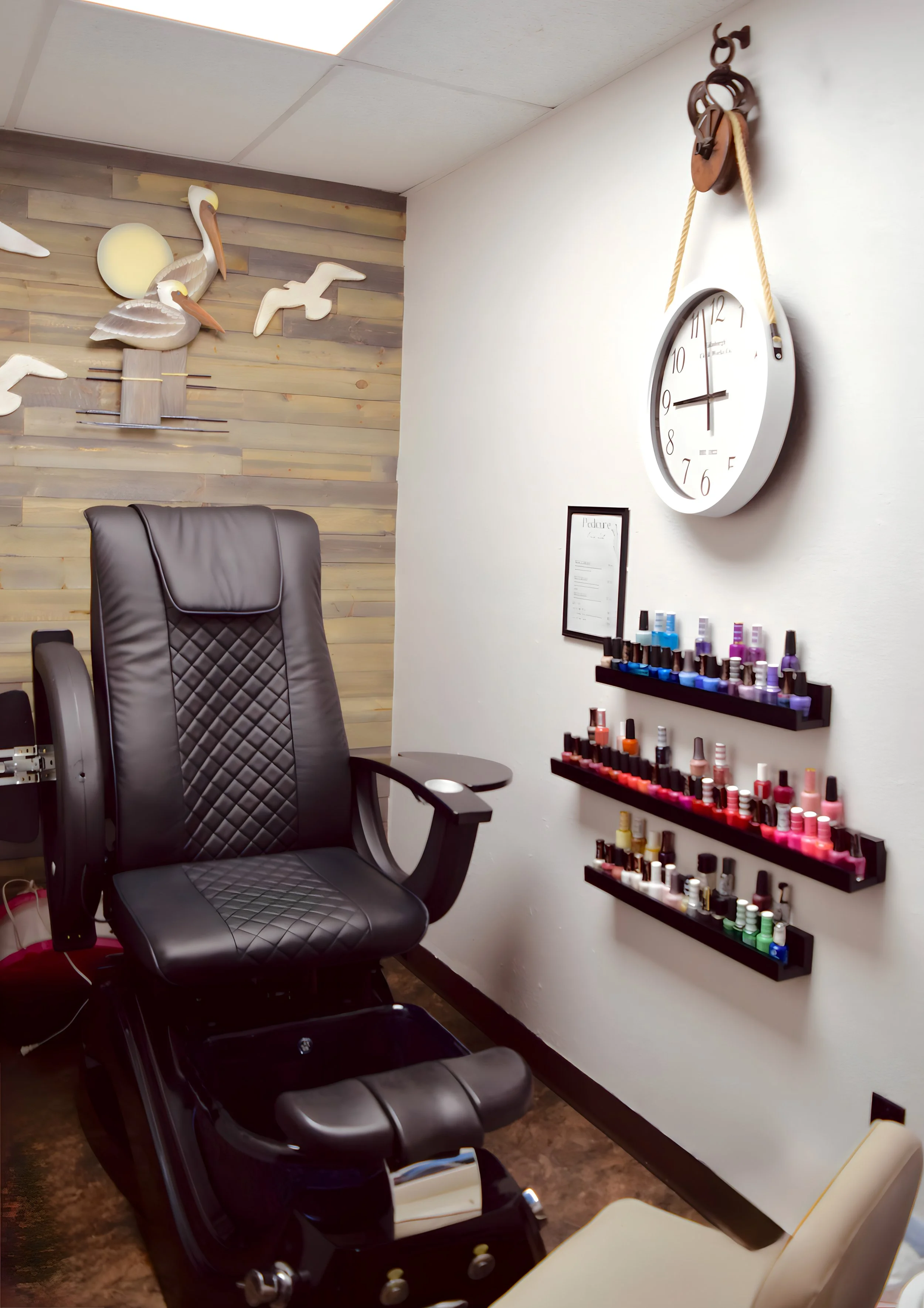 Spa or manicure salon room with a black leather chair, wall-mounted shelves of various nail polish bottles in different colors, a clock, and decorative wall art of birds on a wooden accent wall.