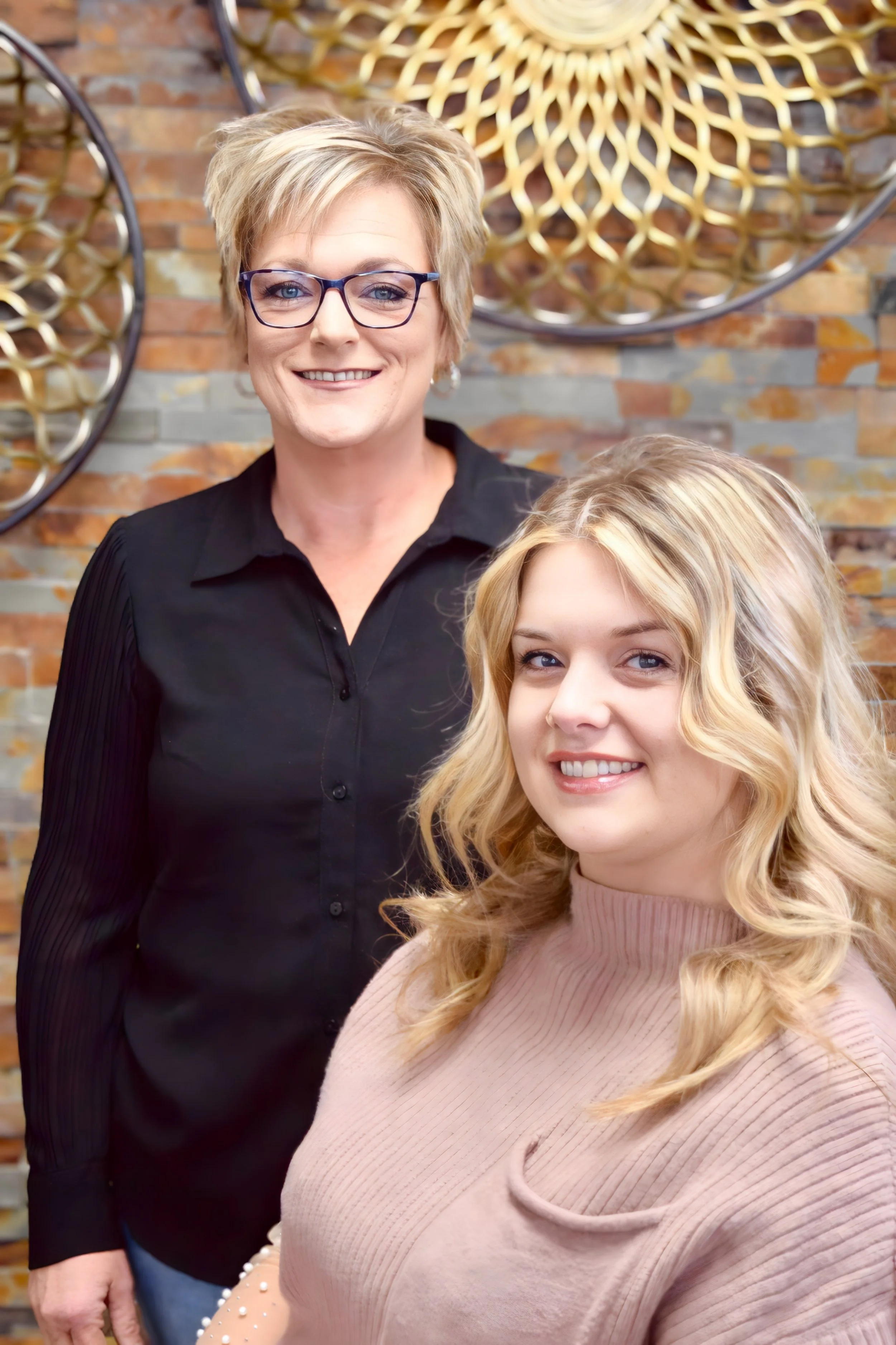 A woman with short blond hair and glasses standing behind a young woman with long wavy blond hair, in front of a brick wall with decorative circular wall art.