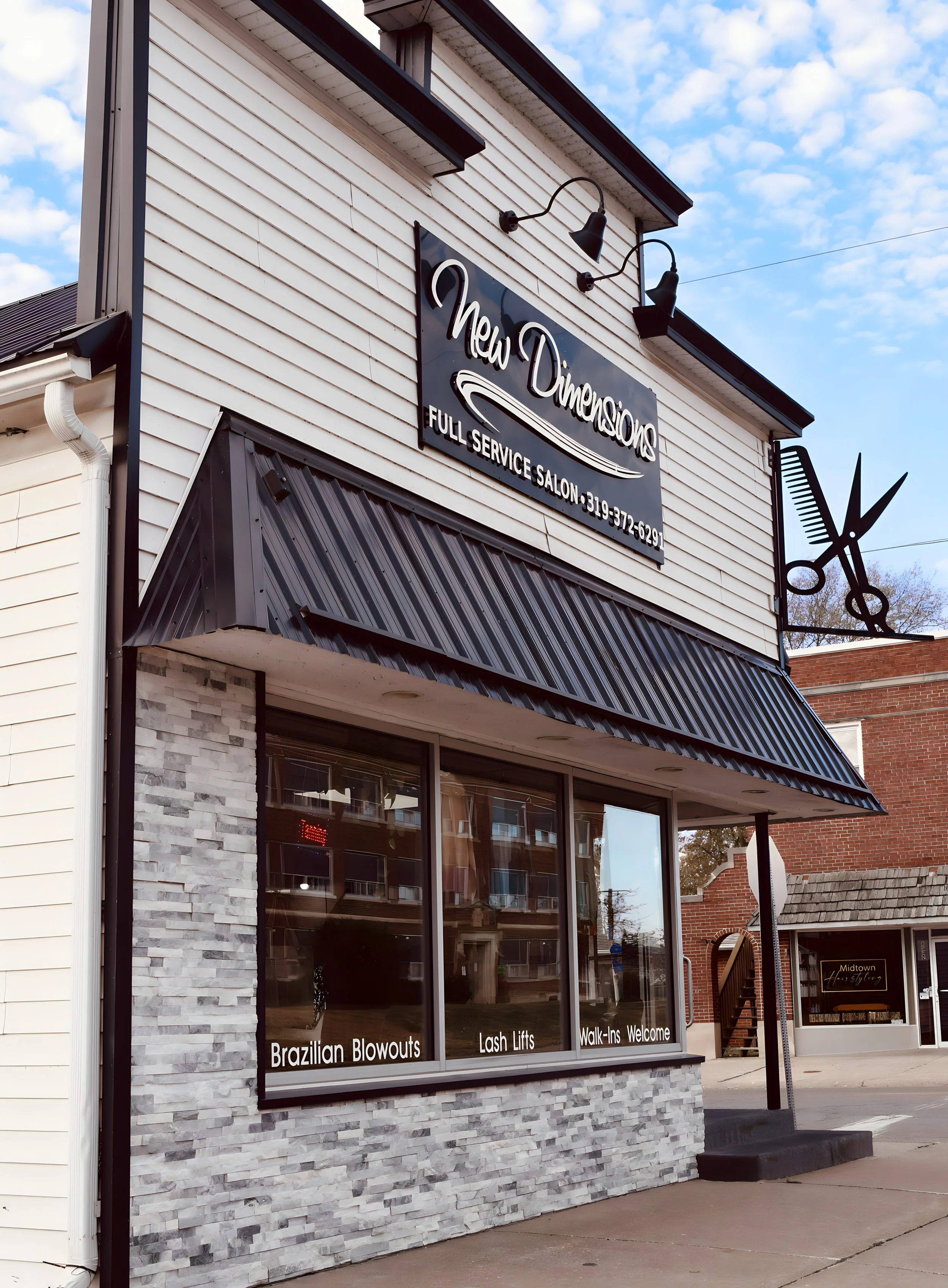 Exterior of a beauty salon named 'New Dimensions' with a large sign, black awning, and decorative scissors sculpture. Window signs advertise Brazilian Blowouts, Lash Lifts, and Walk-Ins Welcome.