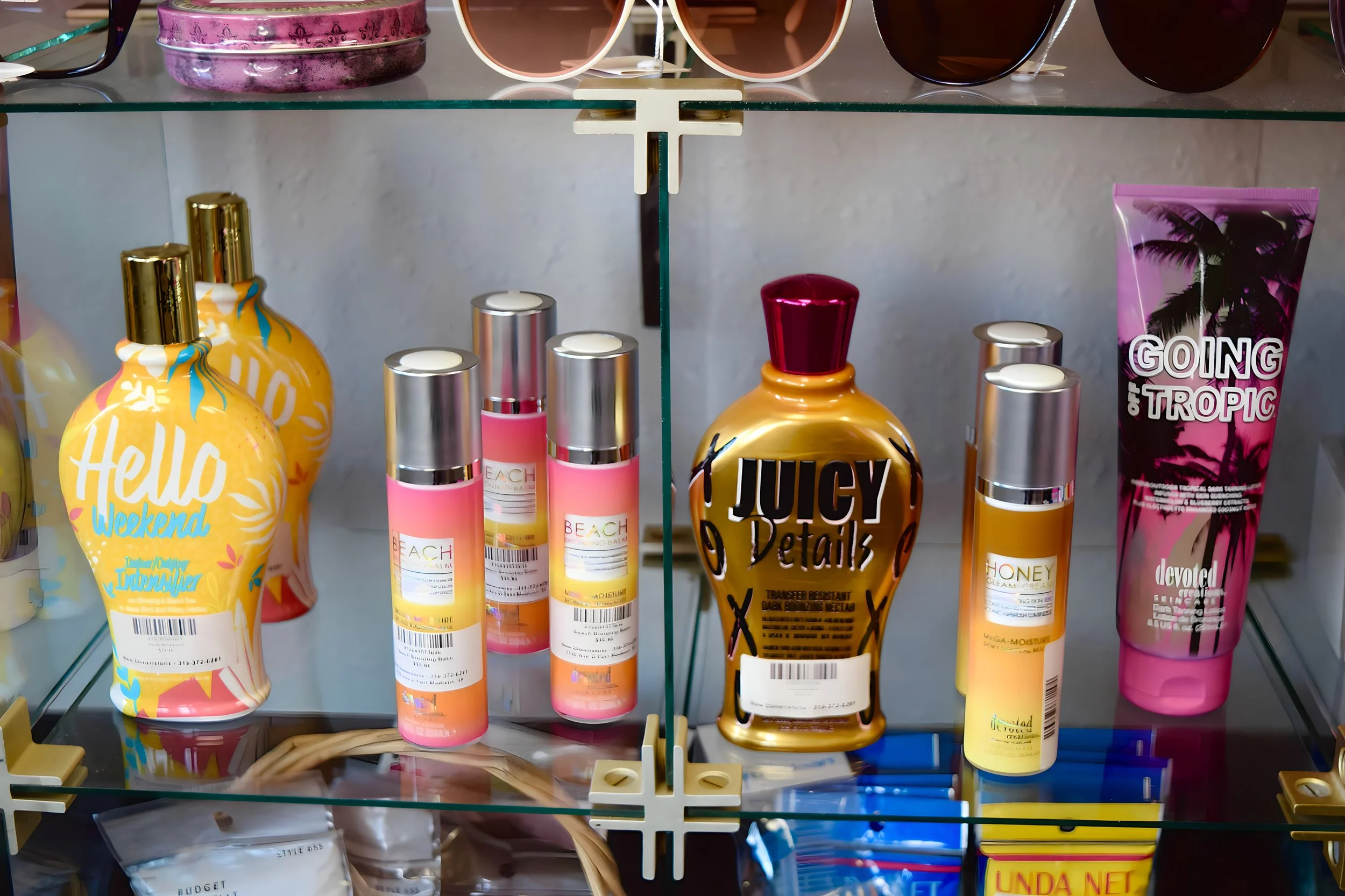 Display shelf with various bottles of skincare and beauty products, including pink and yellow bottles, a large gold bottle labeled 'Juicy Details,' and pink tubes labeled 'Going Tropic' and 'Hello Weekend' with sunglasses on the top shelf.