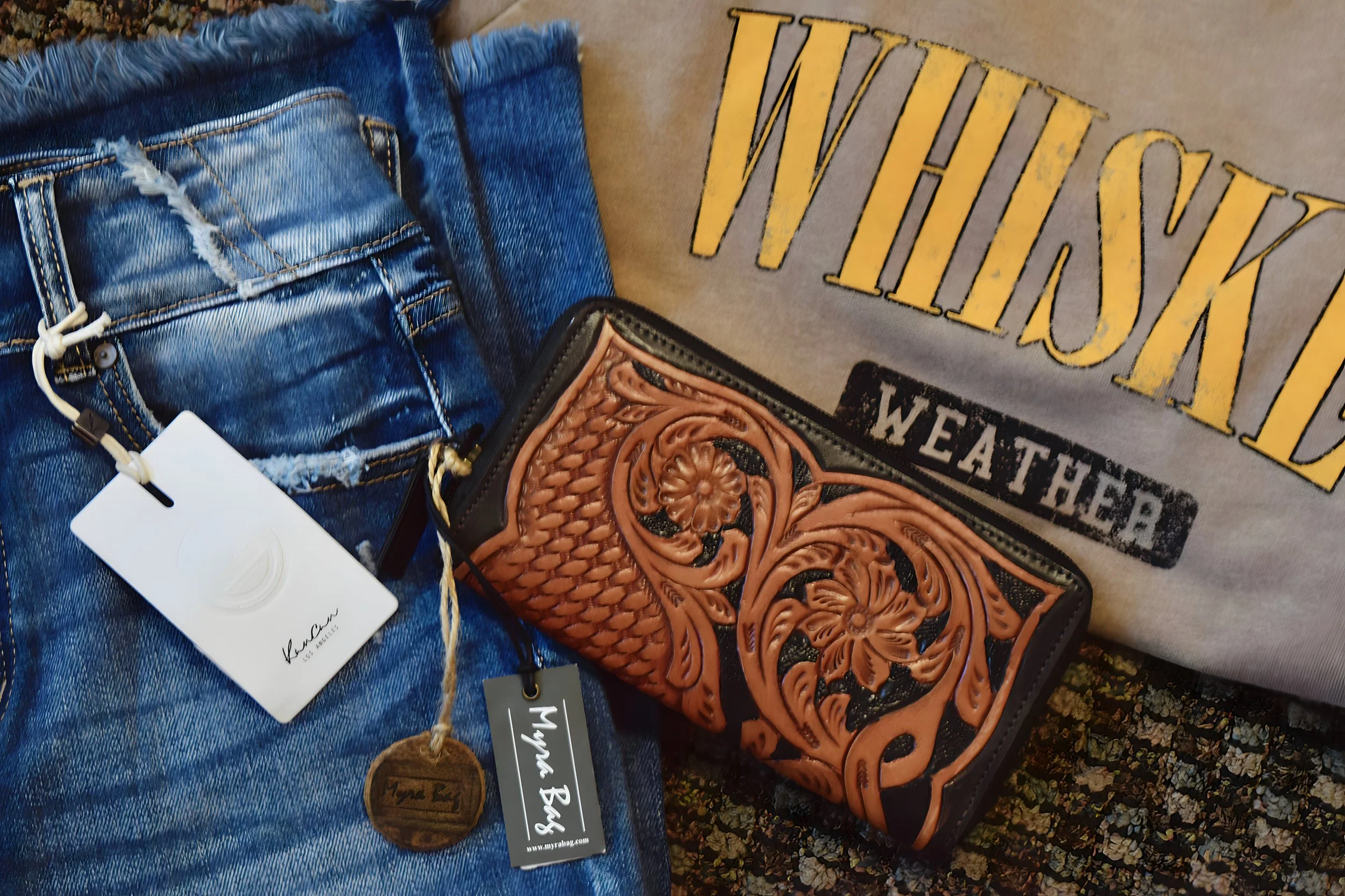 A pair of dark blue distressed denim jeans with a white tag hanging from a loop, a brown leather wallet with embossed floral and swirl patterns, a gray sweatshirt with the words "WISH LIST" in yellow and "WEATHER" in black, and a carpeted floor with 