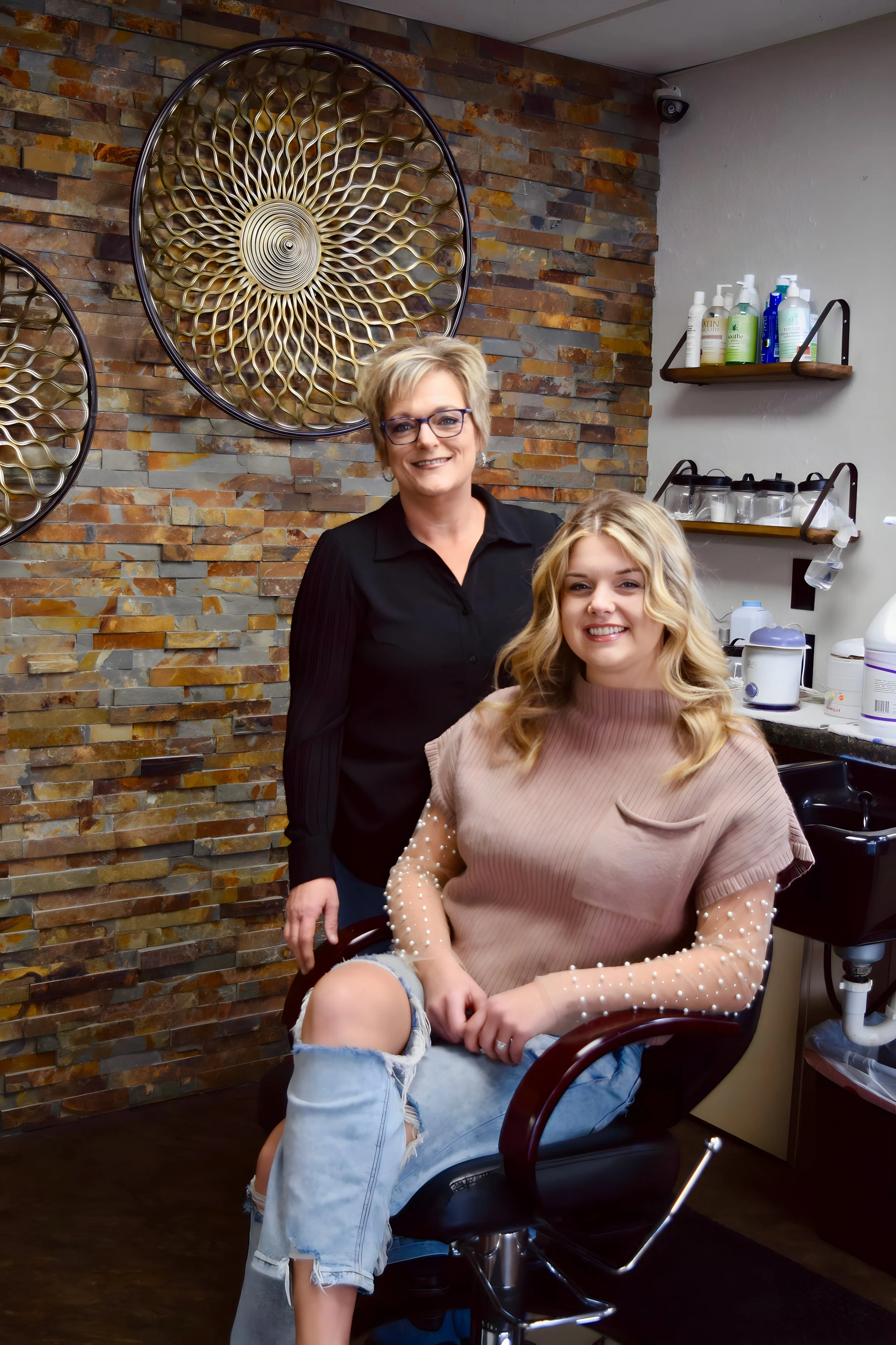 A woman with blonde hair, wearing a beige sweater with pearl embellishments on the sleeves, sitting in a black salon chair at a hair salon, with a woman with short blonde hair and glasses standing behind her. The salon features a brick wall with deco