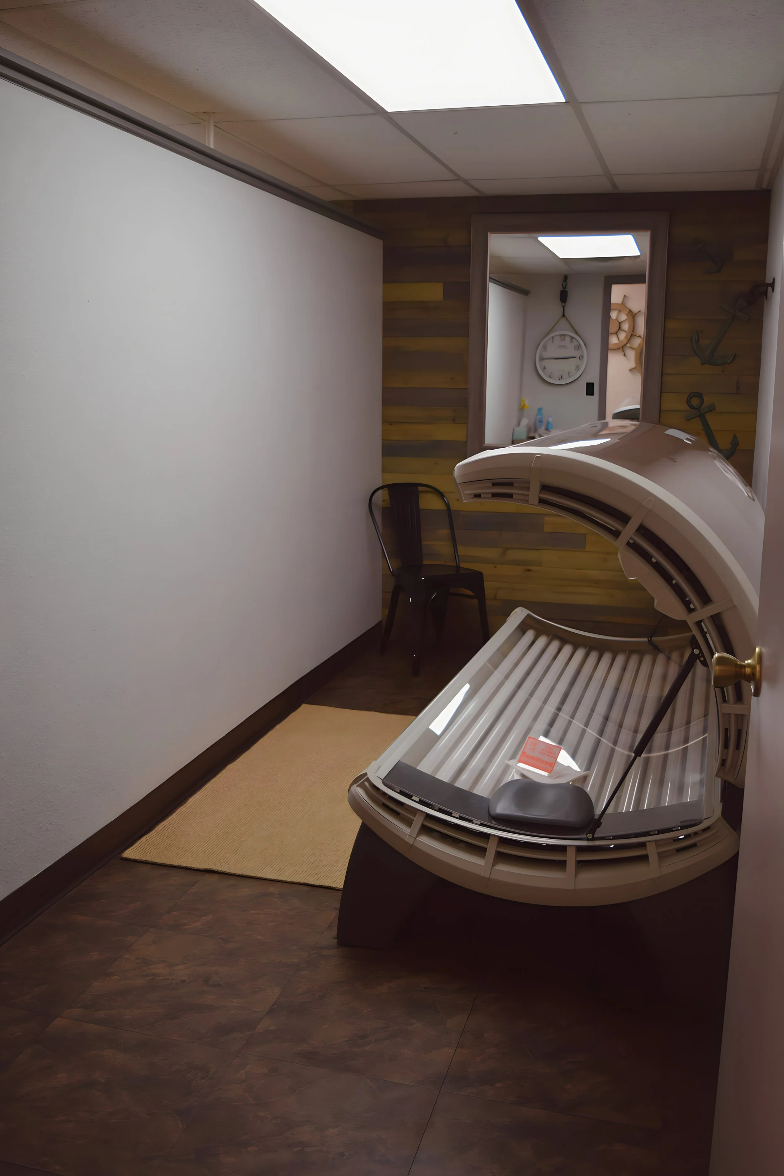 A tanning bed in a small room with dark brown floor tiles, a beige rug, a black chair, and a mirror reflecting a wall clock and nautical decorations.