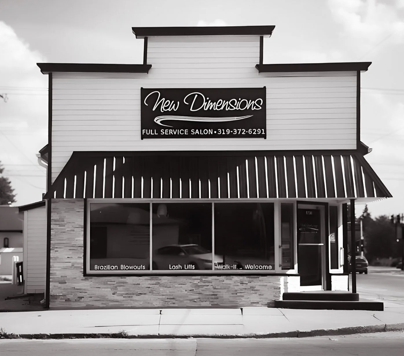 A black and white photo of a 2-story salon building with a large sign that reads "New Dimensions" and a phone number. The ground floor has large windows with signs advertising services like Brazilian blowouts and lash lifts. An awning extends over the sidewalk in front of the building.