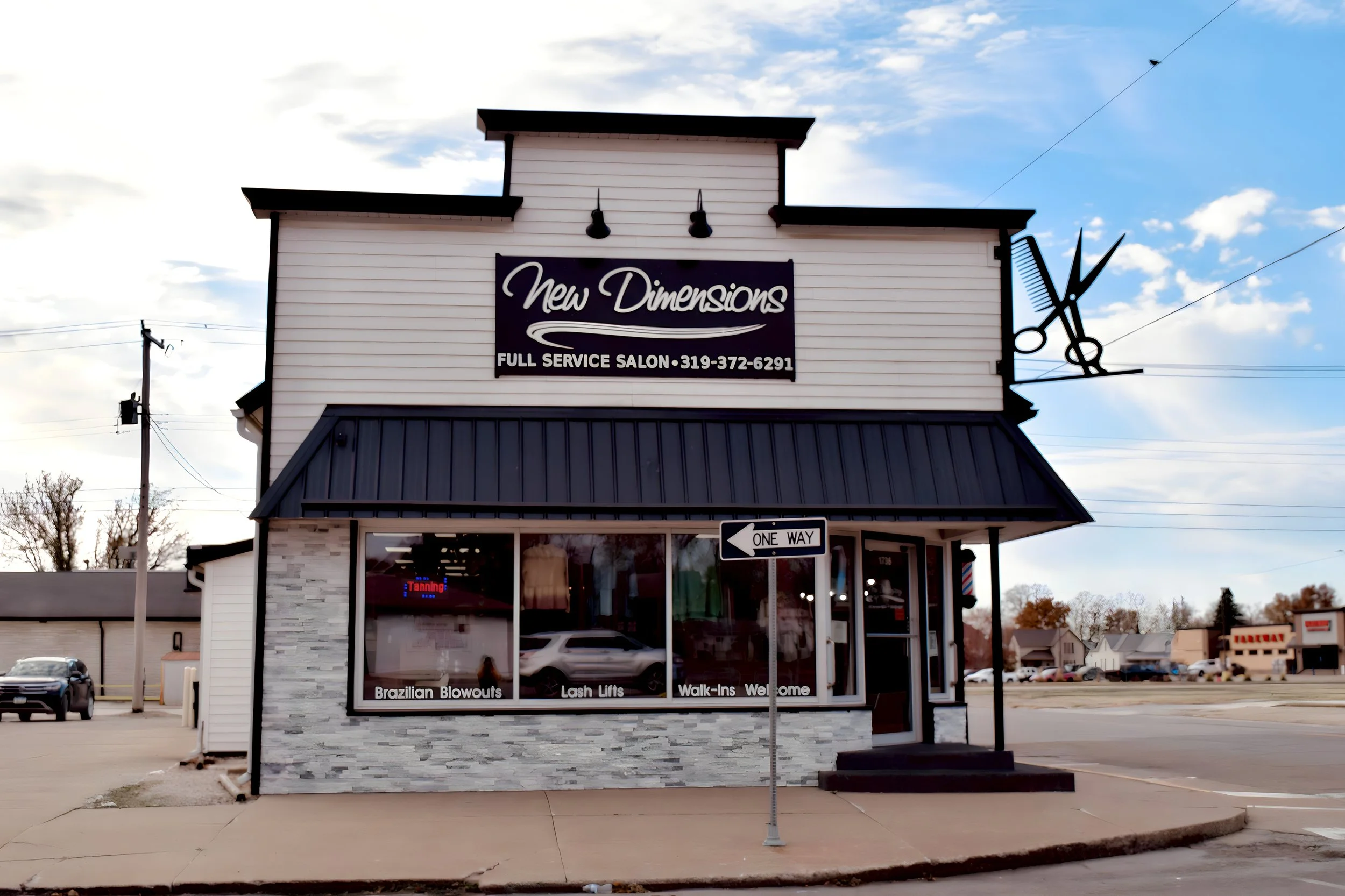 Exterior of a two-story hair salon named 'New Dimensions' with a sign advertising full-service salon, with large front windows displaying services like Brazilian Blowouts, Lash Lifts, and Walk-Ins Welcome. A one-way street sign is in front of the bui