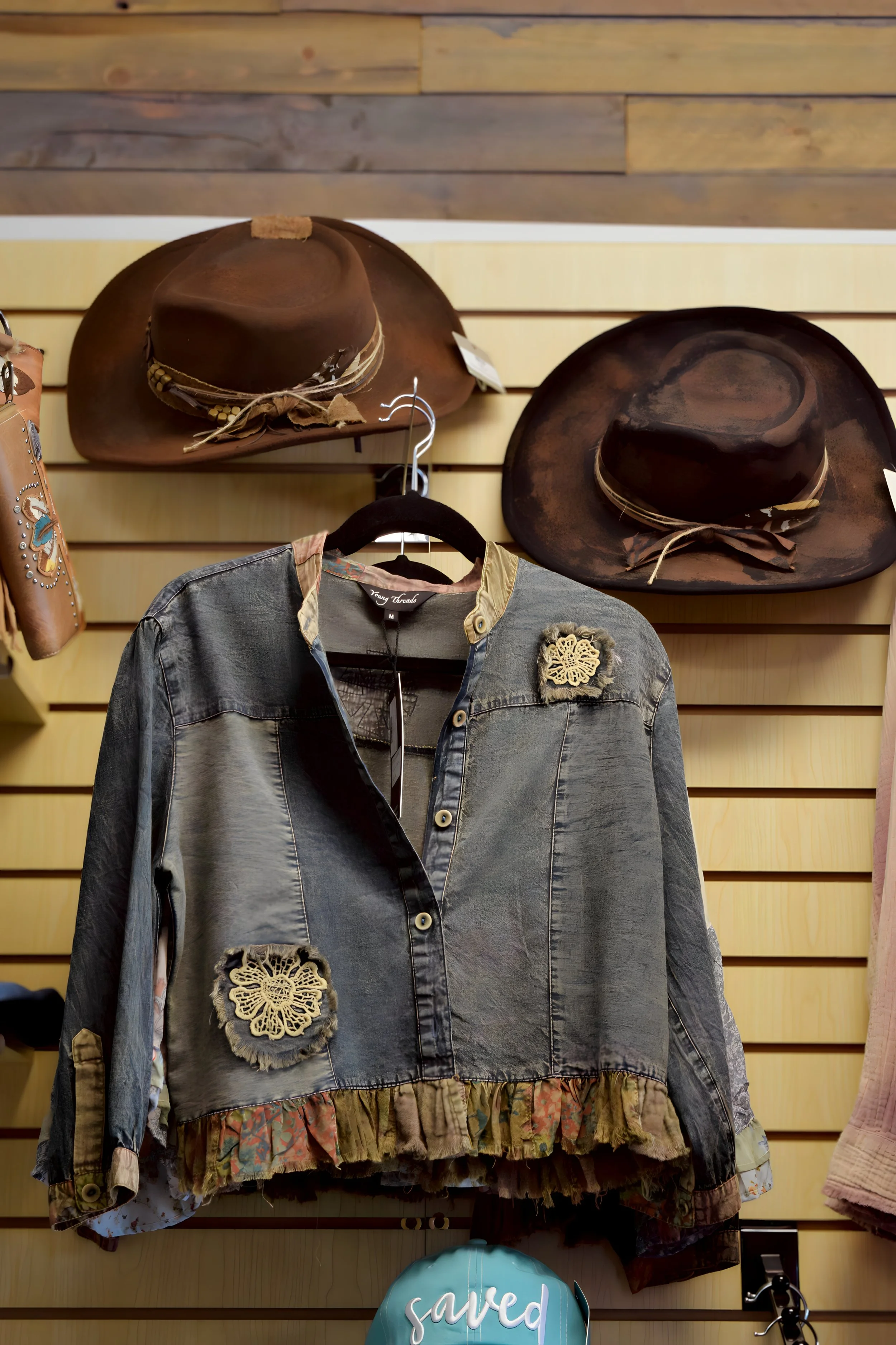 A display of two cowboy hats, a denim jacket with decorative patches on a wooden wall in a store.