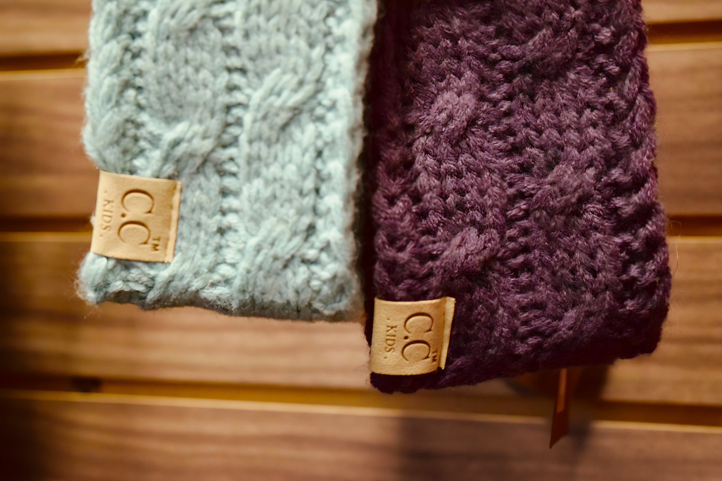 Close-up of two folded knitted hats, one light gray and one dark purple, with leather tags reading "CO KIDS" attached to each hat.