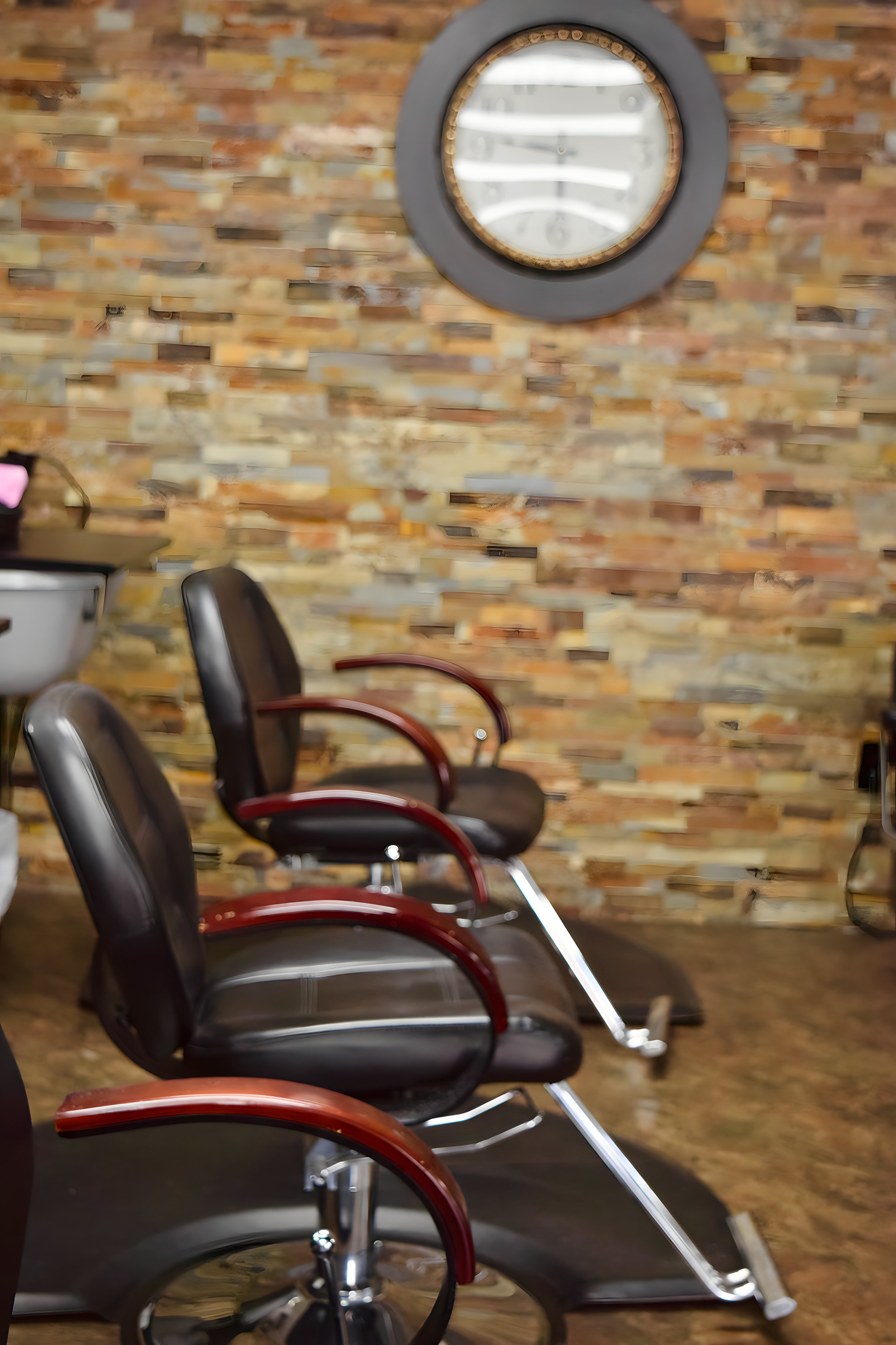 Two black salon chairs with wooden armrests in front of a brick wall, with a round mirror hanging on the wall.